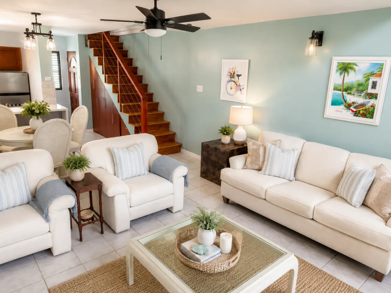 Living room with white sofas, a glass coffee table, and a staircase in the background. The walls are light blue.