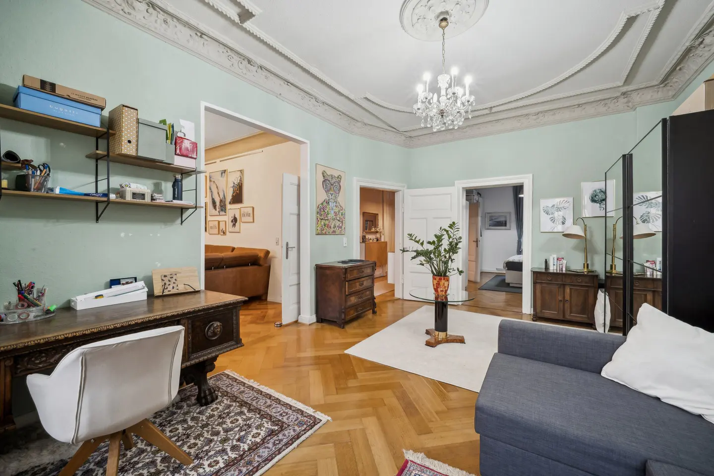 A living room with light green walls, hardwood floors, and ornate ceiling trim. A desk, sofa, and doorways to other rooms are visible.