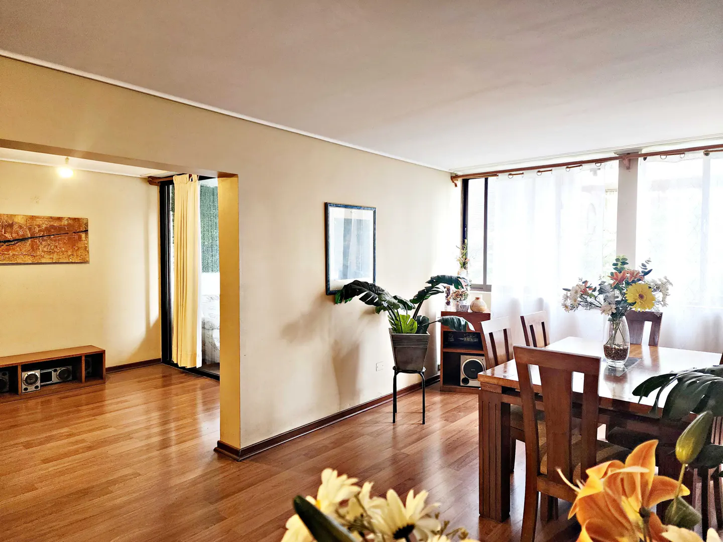 Open-concept living and dining area with hardwood floors, beige walls, and natural light. Table with flowers, plants, and artwork.