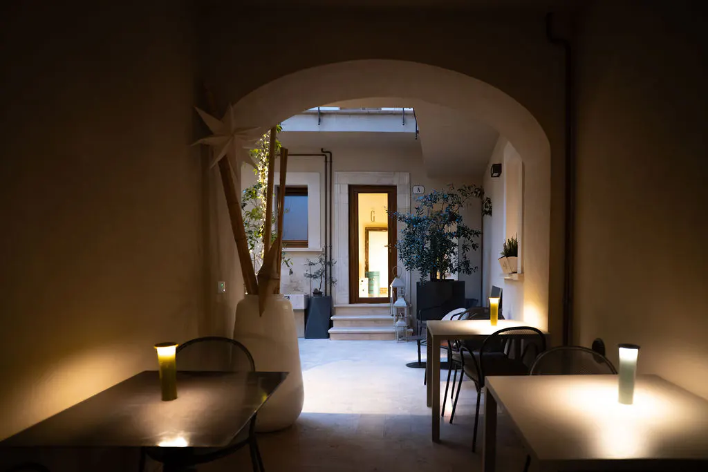 Indoor patio with tables and chairs under an archway leading to an outdoor courtyard with plants and a doorway.