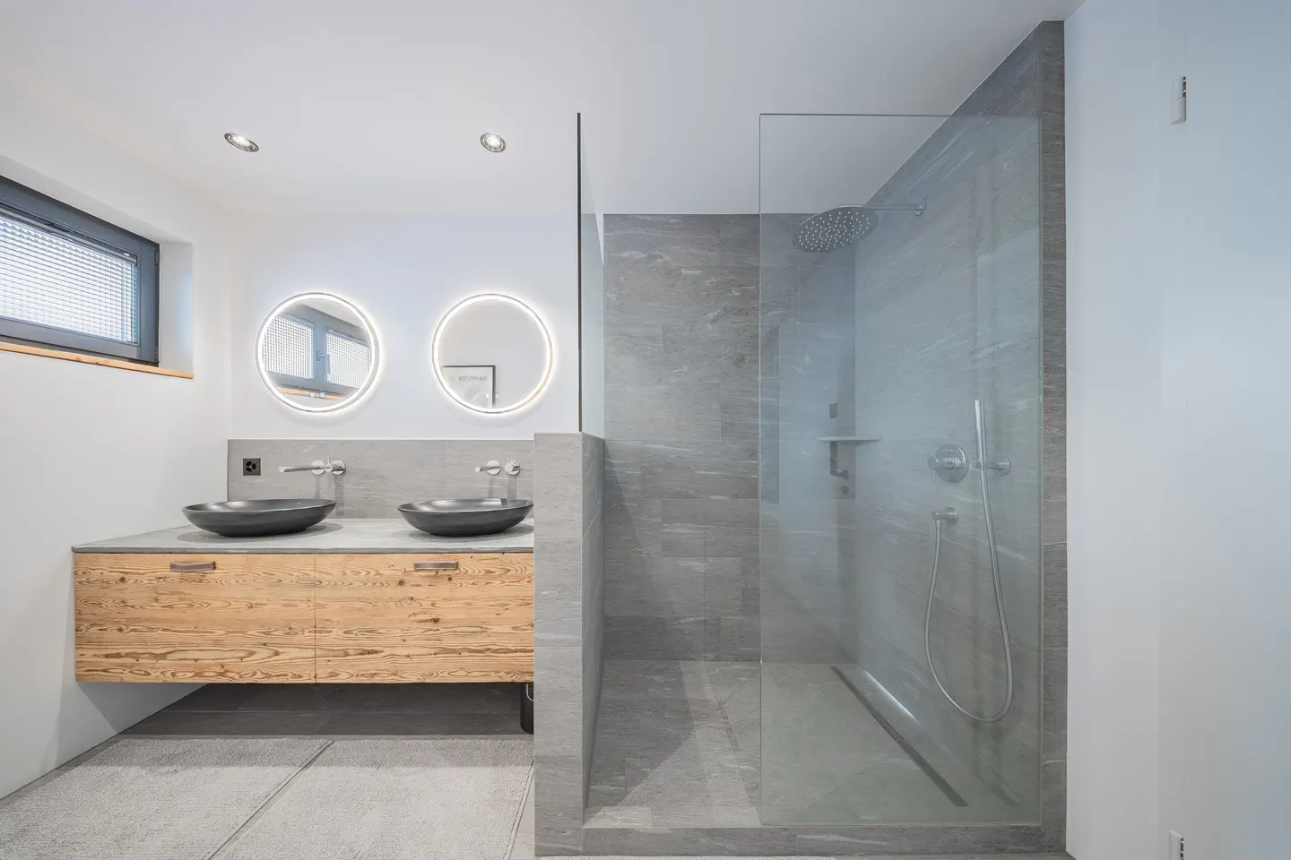 Modern bathroom with a glass shower, gray tile, and a wood vanity with two black vessel sinks and round, lighted mirrors.