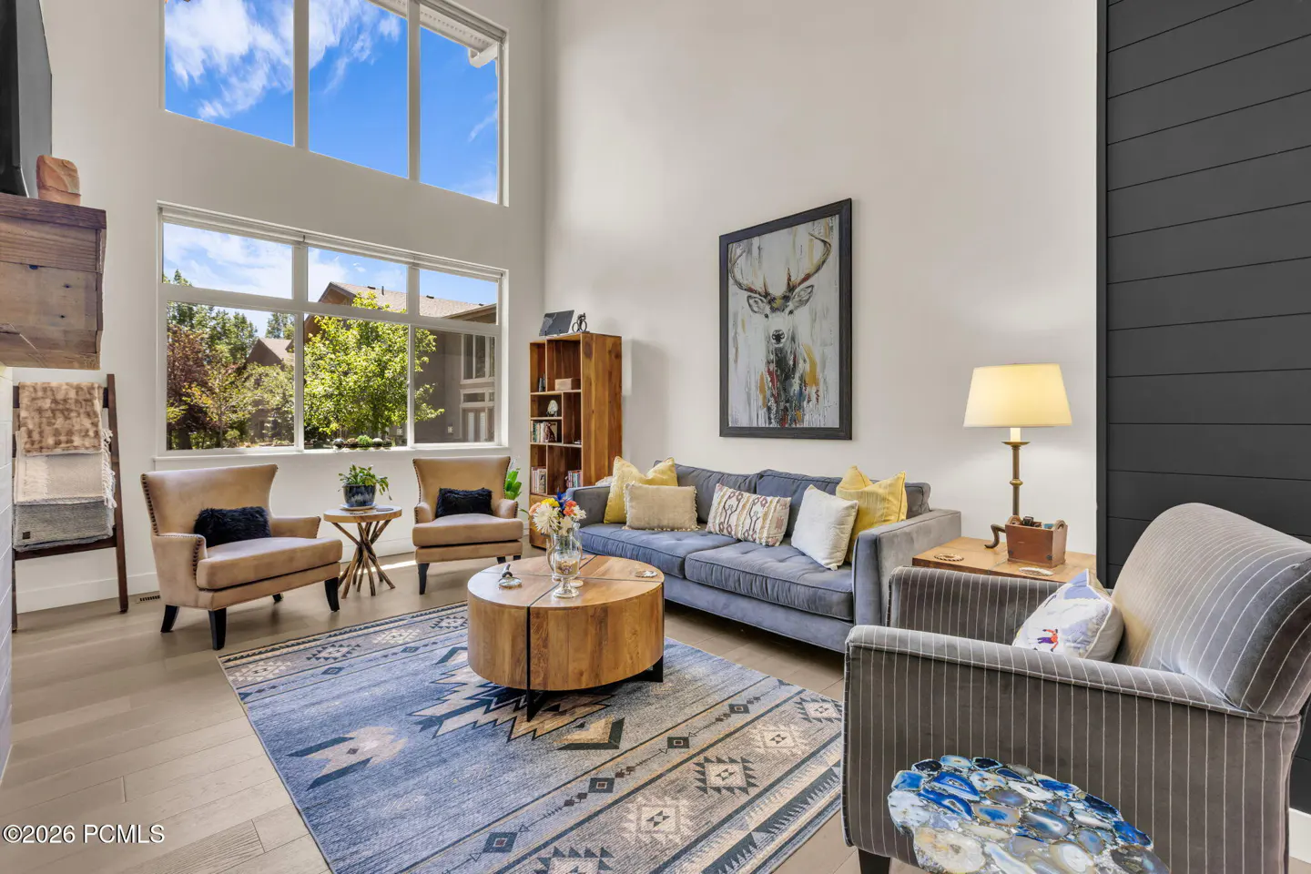Living room with high ceilings, large windows, grey sofa, two tan armchairs, round wood coffee table, and patterned rug.