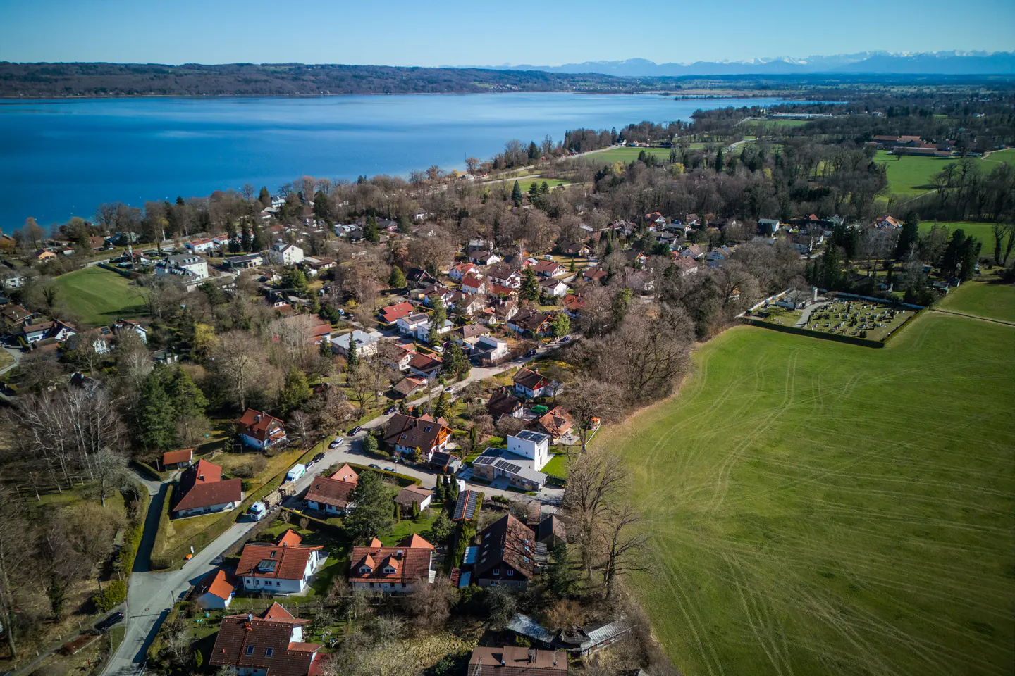Aerial view of a village nestled between a large blue lake and green fields, with distant mountains under a clear sky.