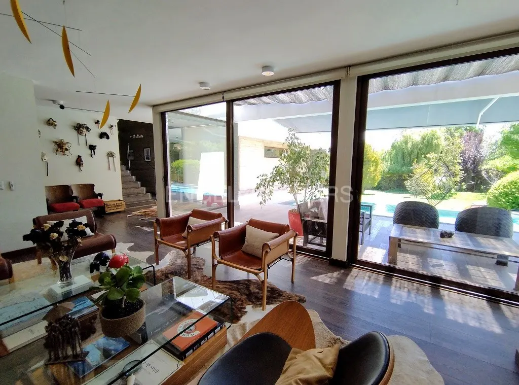 Living room with dark wood floors, brown leather chairs, and a glass coffee table. Large windows overlook a pool and green backyard.