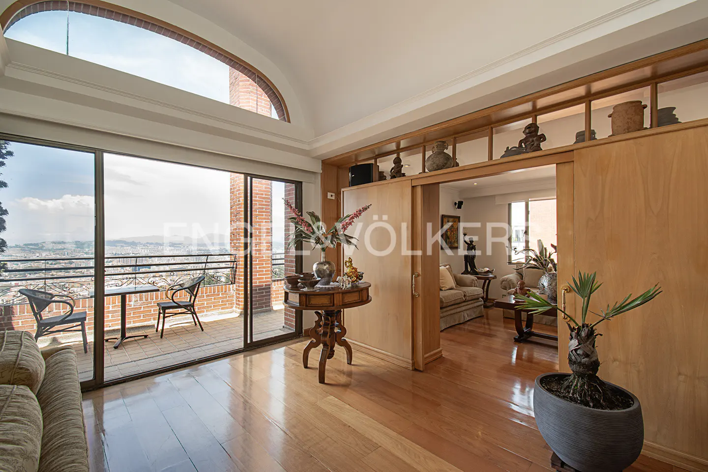 A bright living room with hardwood floors, a balcony with a city view, and a potted plant in the foreground.