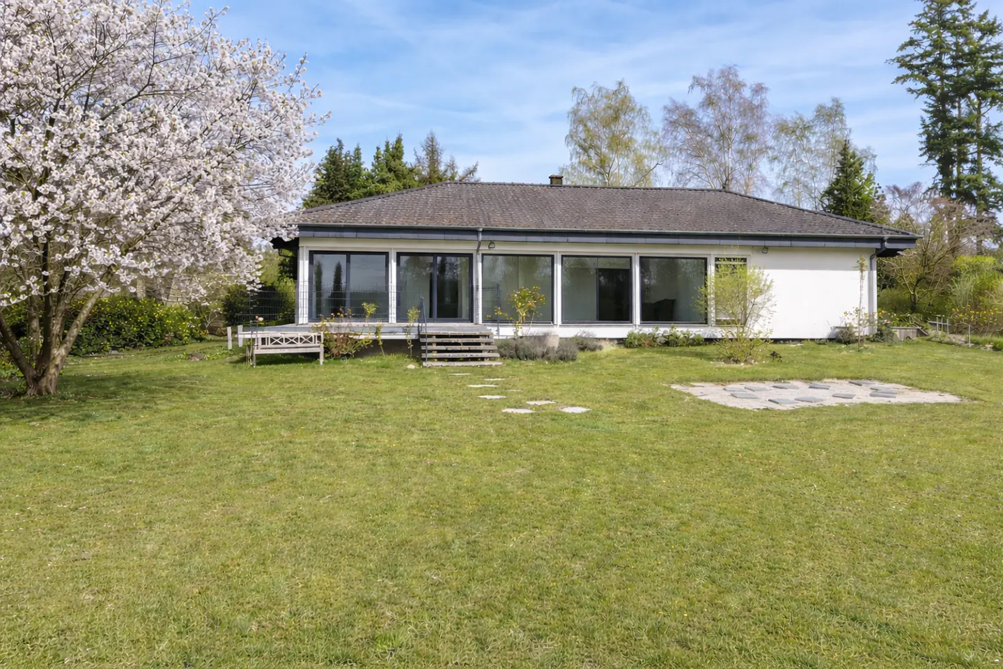 A modern, one-story white house with large windows and a gray roof is set on a green lawn with trees.