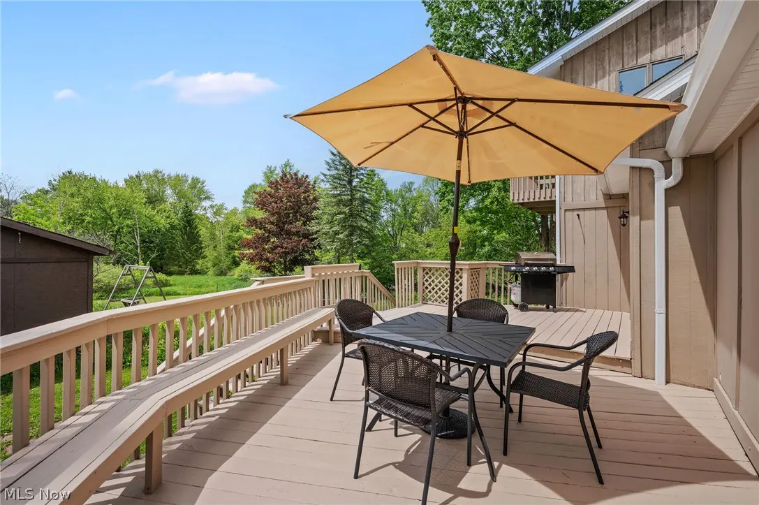 Wooden deck with a black dining set, tan umbrella, and grill, overlooking a lush green backyard on a sunny day.