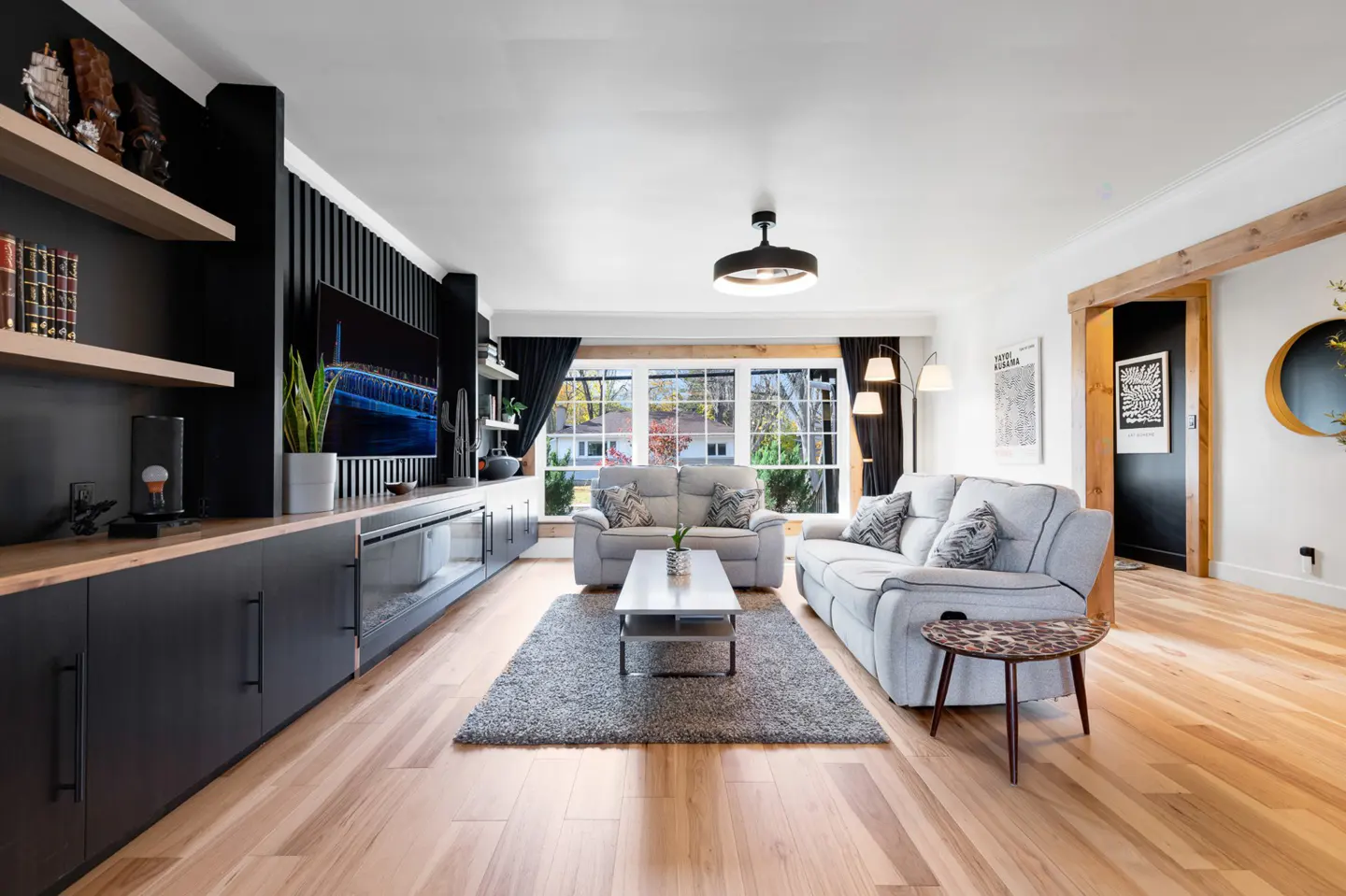 Bright living room with hardwood floors, gray sofas, and a large window. Black built-in cabinets and shelves line one wall.