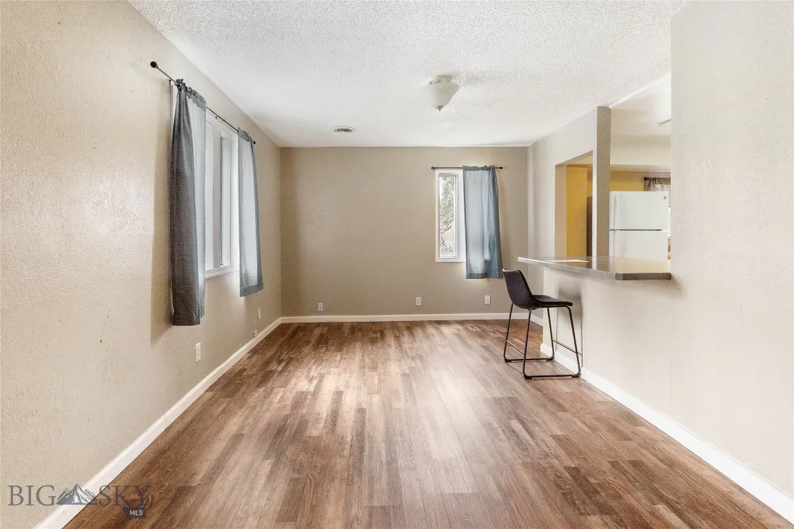 A bright, empty living room with wood floors, beige walls, and gray curtains. A black stool sits near a kitchen counter.
