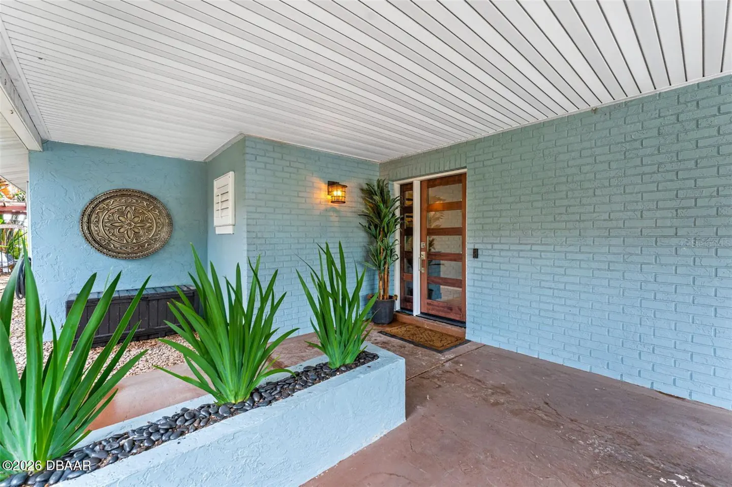 Covered porch with blue brick walls, a modern wood door, and a planter with green plants and black rocks.