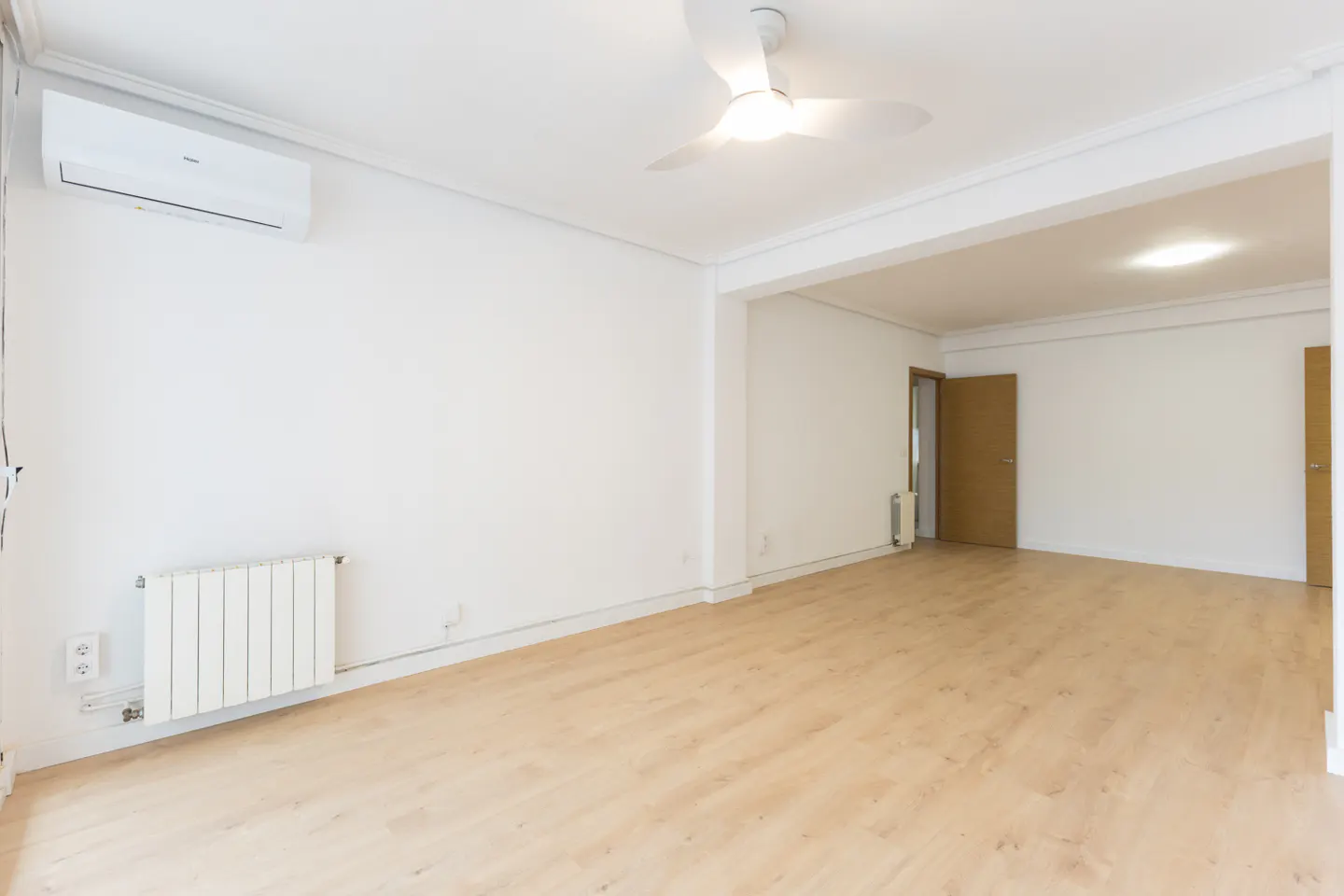 Bright, empty room with white walls, light wood floors, a ceiling fan, wall-mounted AC unit, and a white radiator.