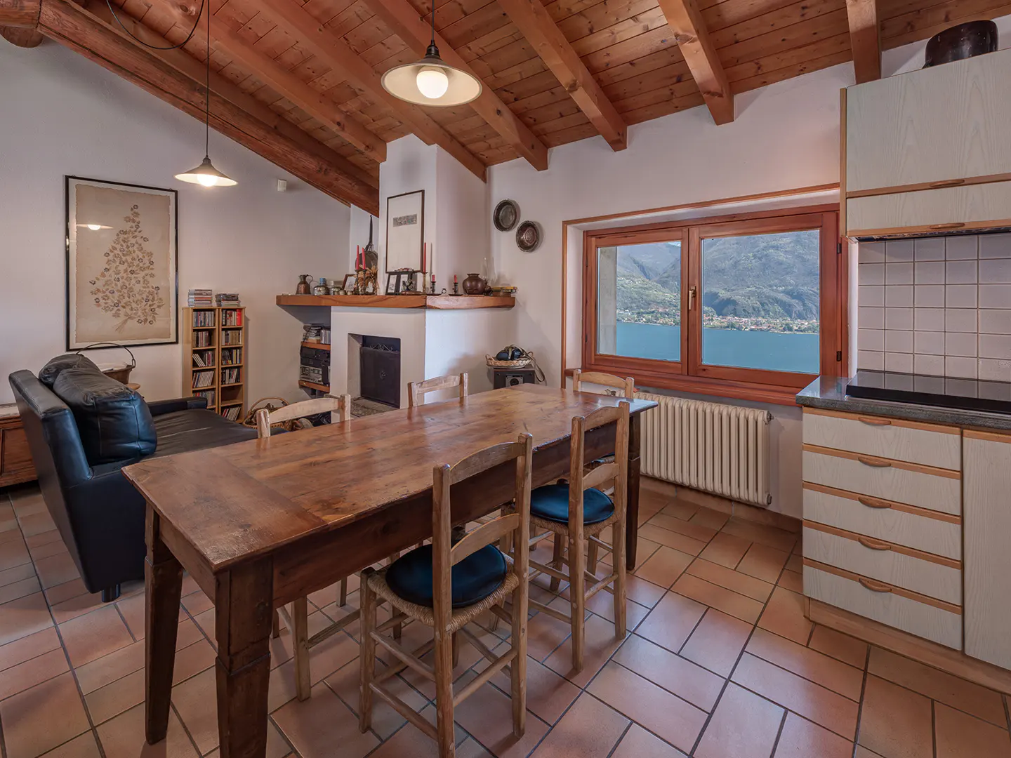 A bright living space with a wood table, chairs, and a view of a lake through a window. The room has a fireplace, a couch, and a wood-paneled ceiling.
