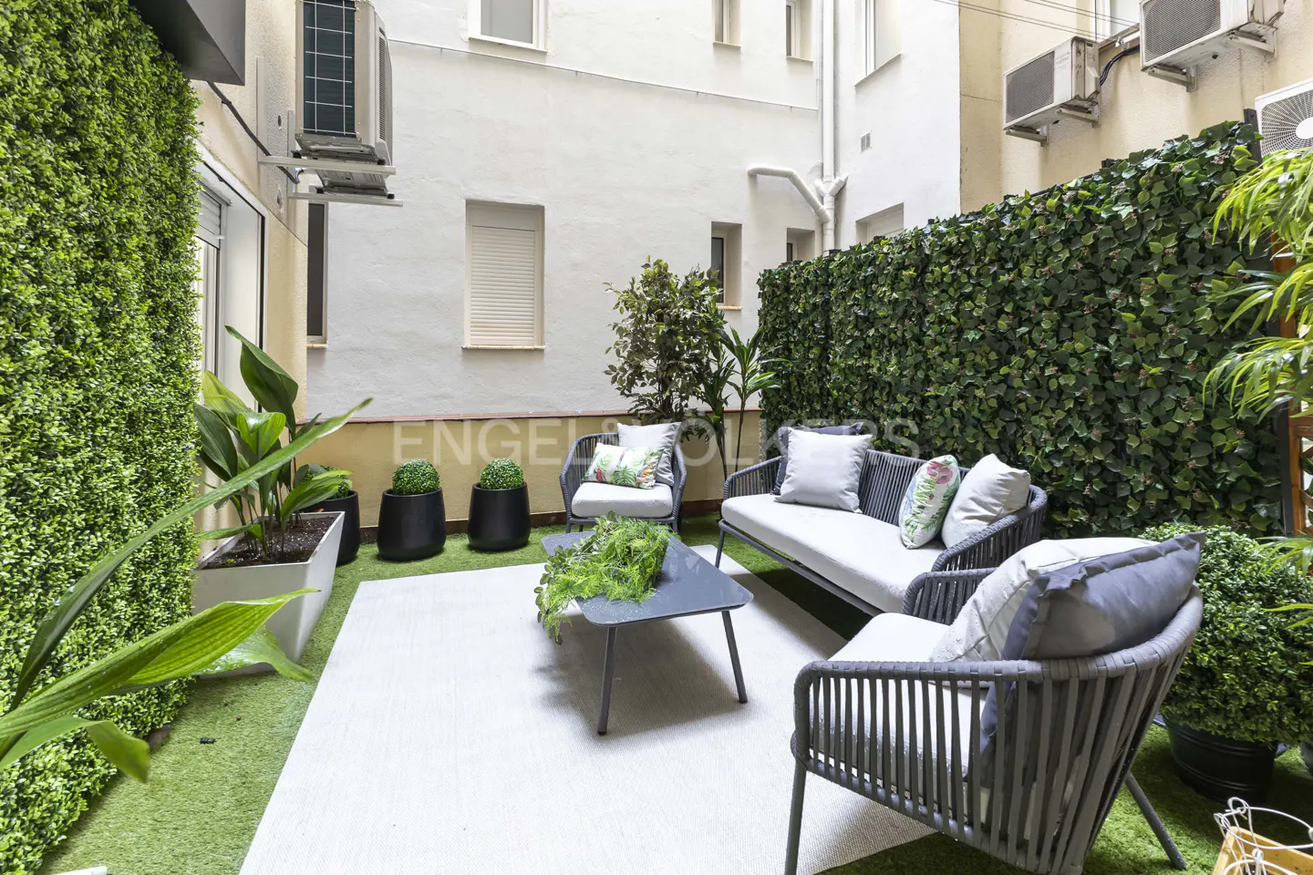 Outdoor patio with gray wicker furniture, white cushions, and green foliage wall. A gray coffee table sits on a white rug.