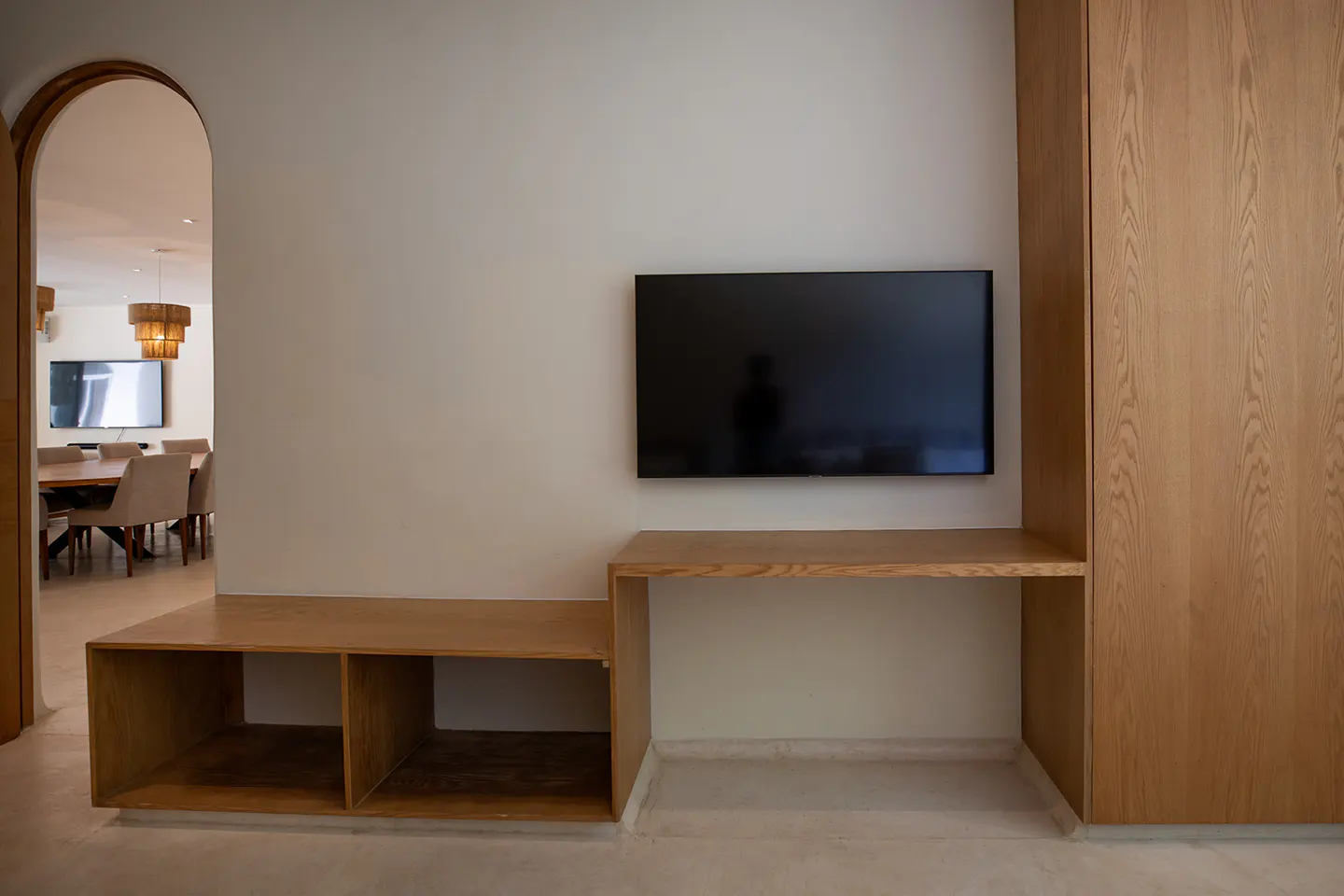 Wall-mounted TV above a custom light wood desk and storage unit, with an arched doorway leading to a dining area.
