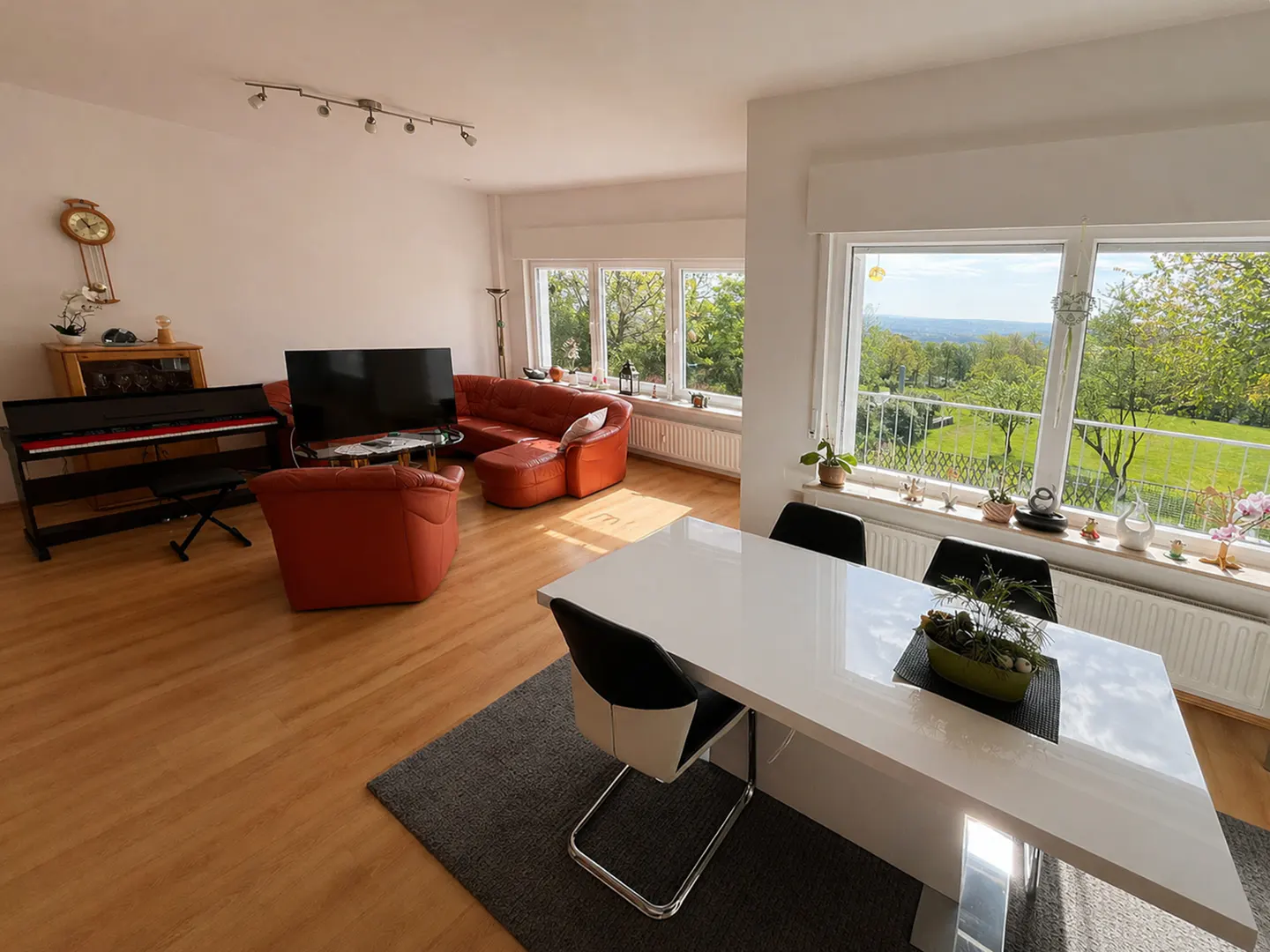 Bright living room with a white dining table, red leather sofa, piano, and large windows overlooking a green landscape.