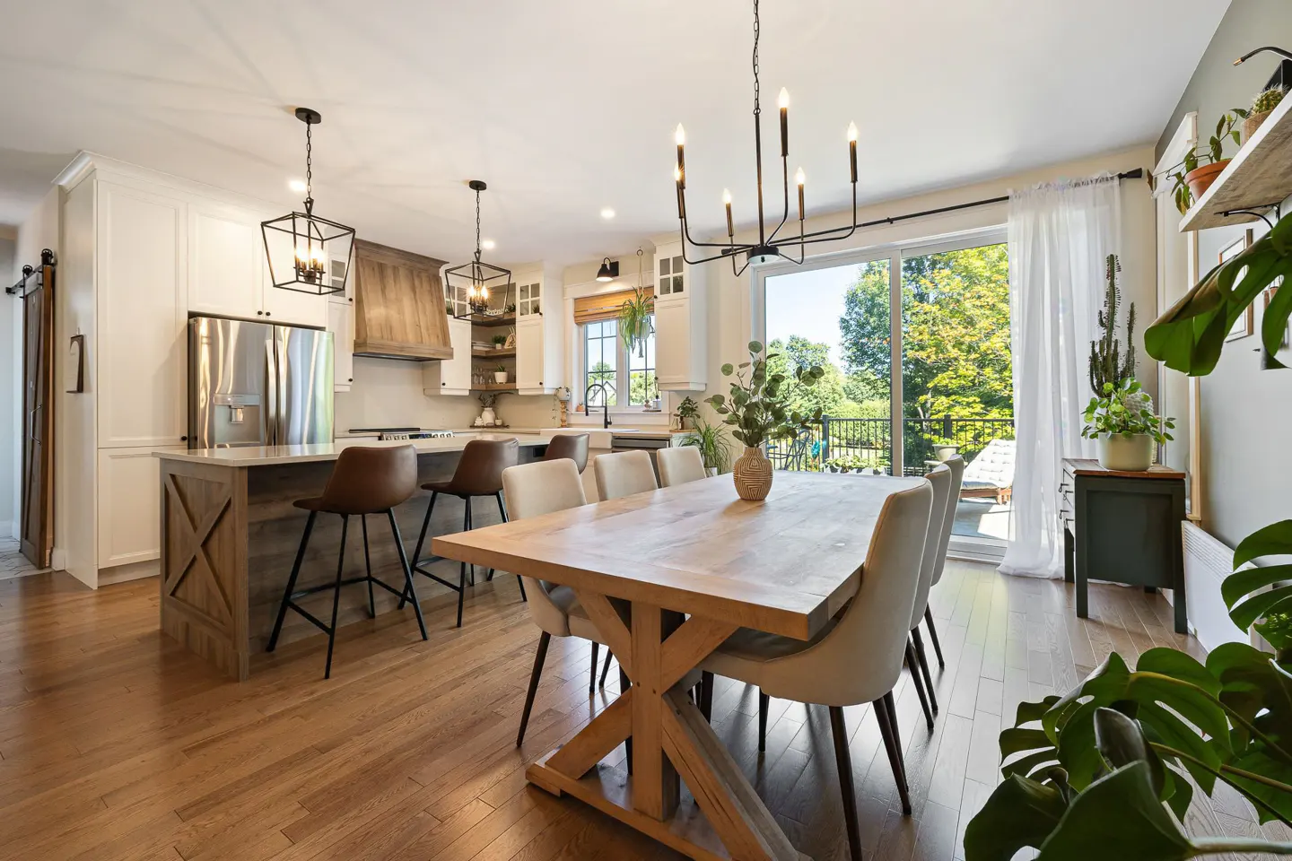 Open-concept kitchen and dining area with wood floors. A large wooden table with beige chairs sits under a black chandelier. Sliding glass doors lead to a balcony.