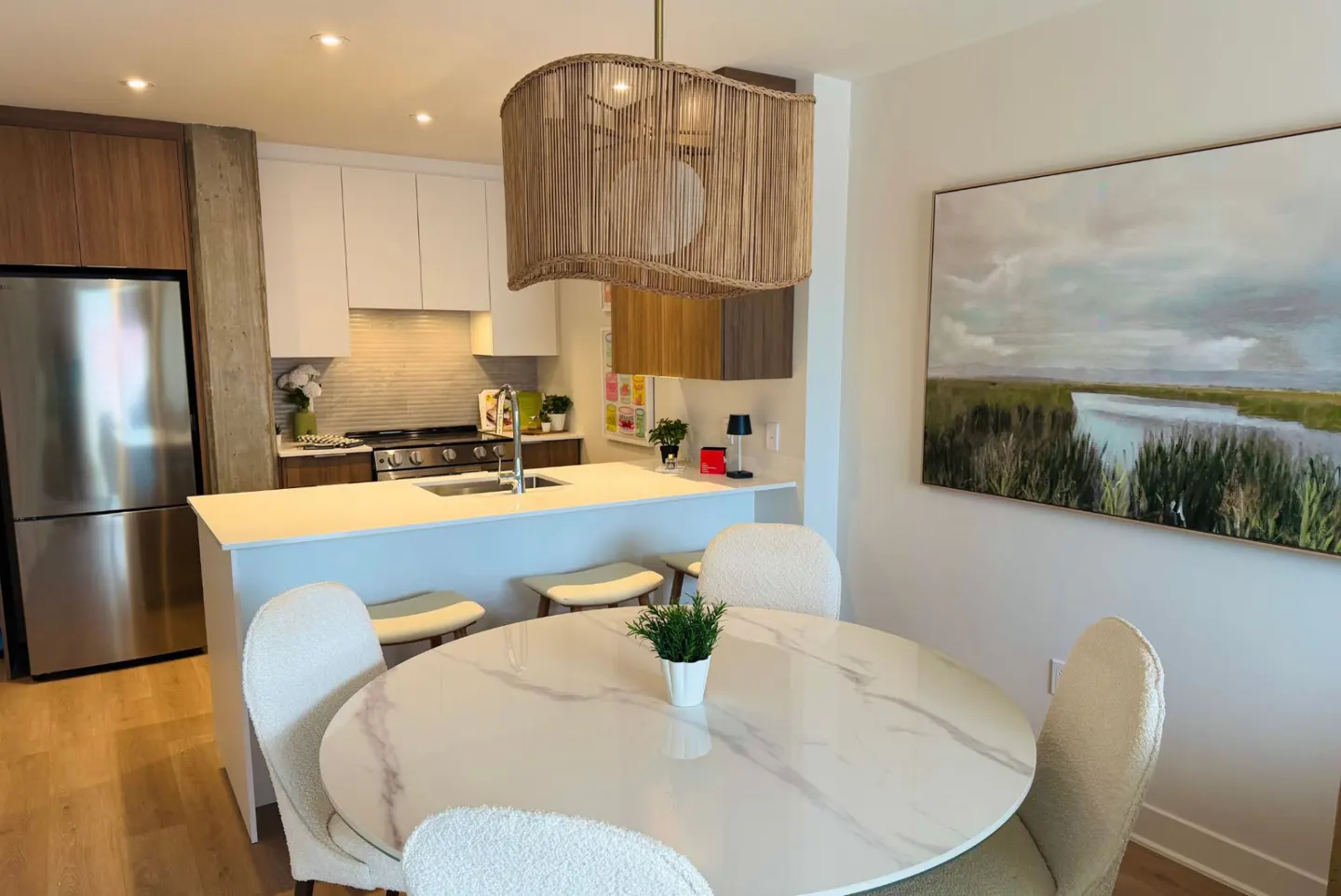 A modern kitchen and dining area with a marble table, white chairs, and a woven pendant light. Stainless steel appliances and a landscape painting are visible.