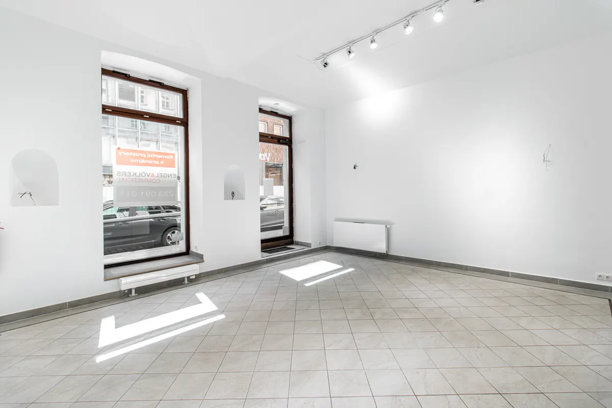 Bright, empty room with white walls, tile floor, and track lighting. Windows and a door show a street scene outside.