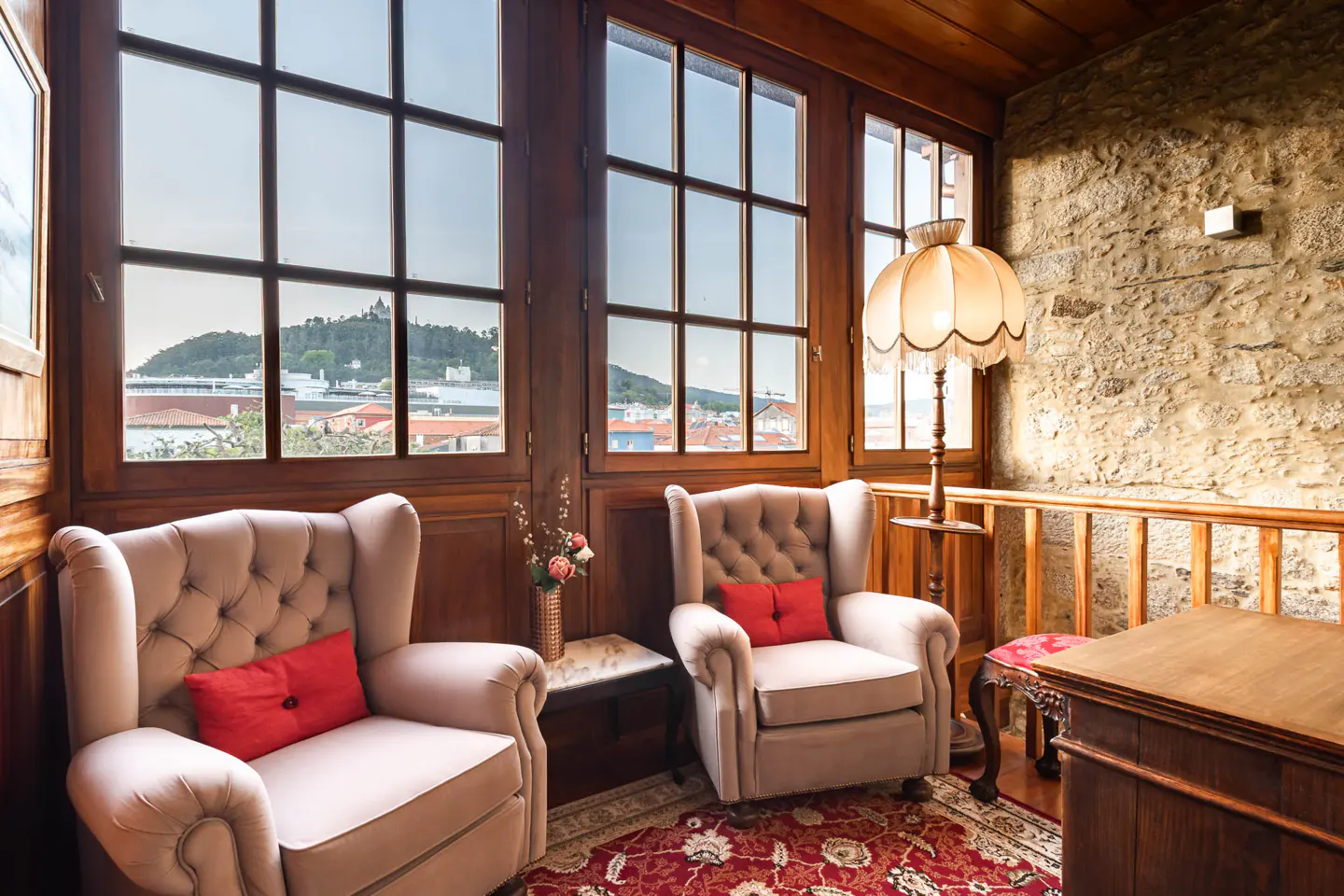 Cozy room with two tufted armchairs, red pillows, and large windows overlooking a cityscape. A vintage lamp and stone wall add character.