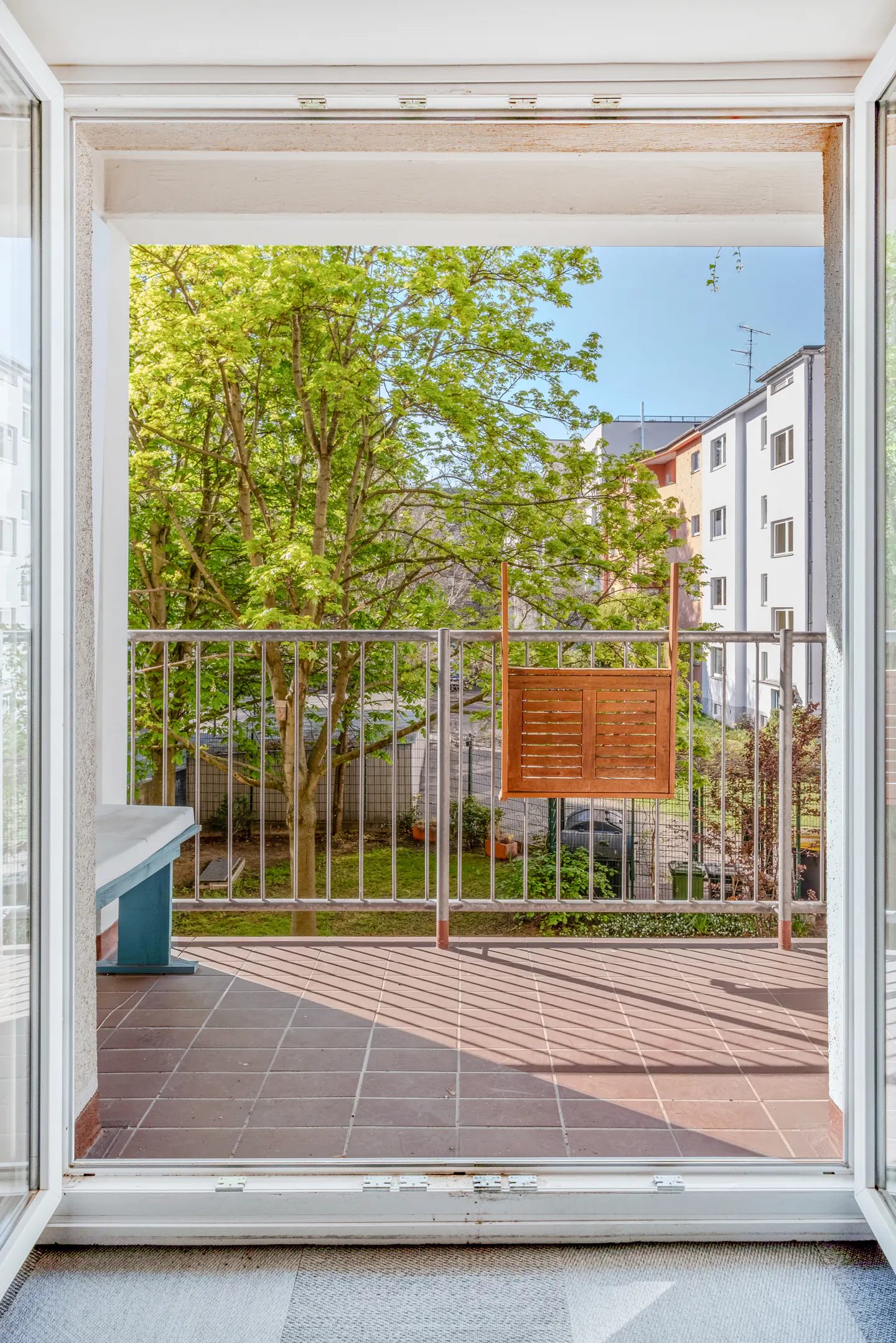 View from open patio doors onto a tiled balcony with metal railing, overlooking green trees and buildings.