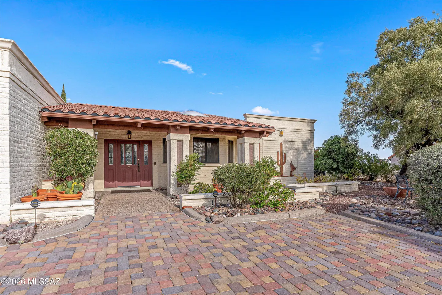 Front view of a single-story home with a red tile roof, brick driveway, and desert landscaping under a blue sky.