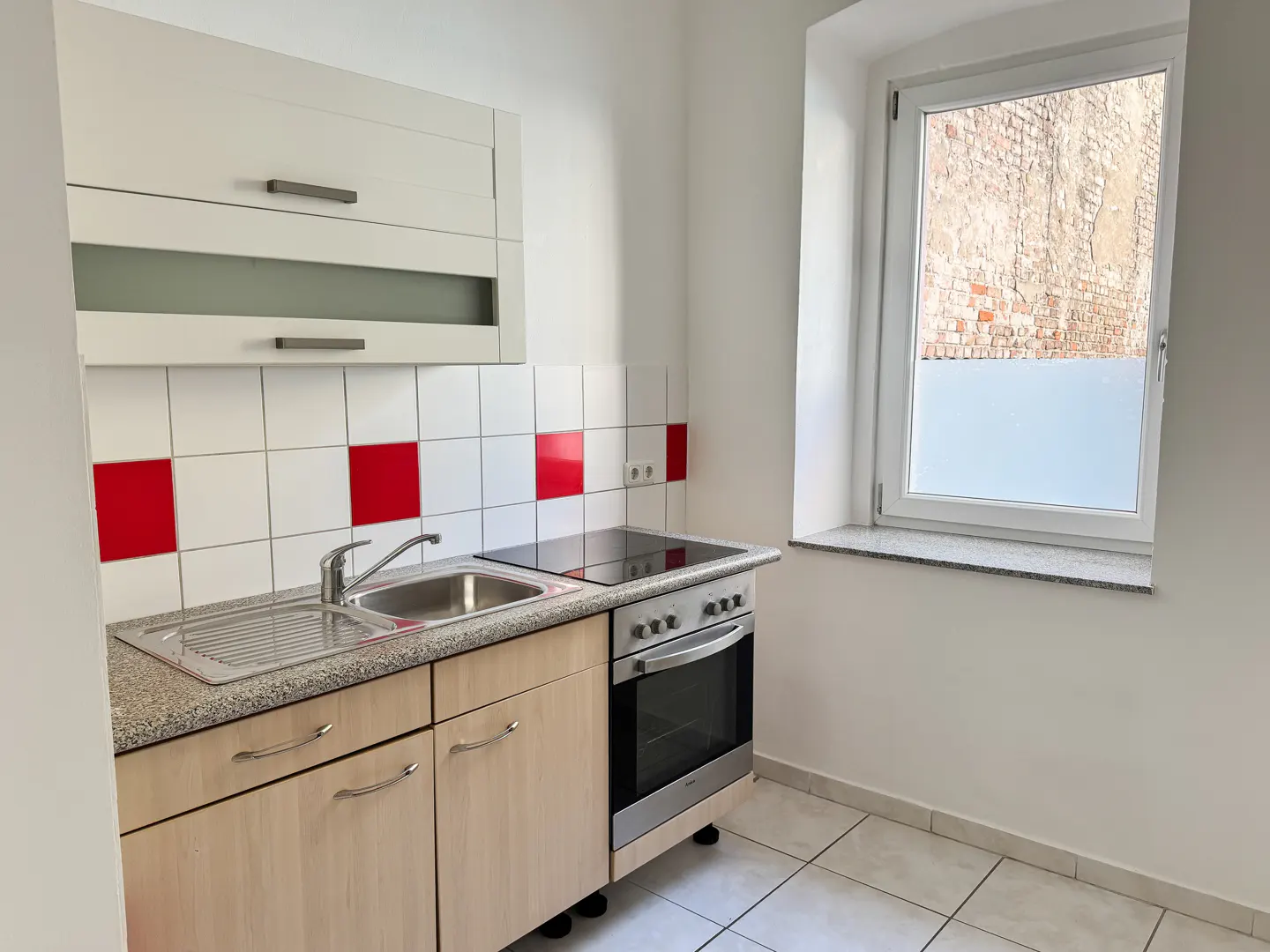A small kitchen with light wood cabinets, a stainless steel sink, and an oven. White and red tiles line the wall. A window shows a brick building.