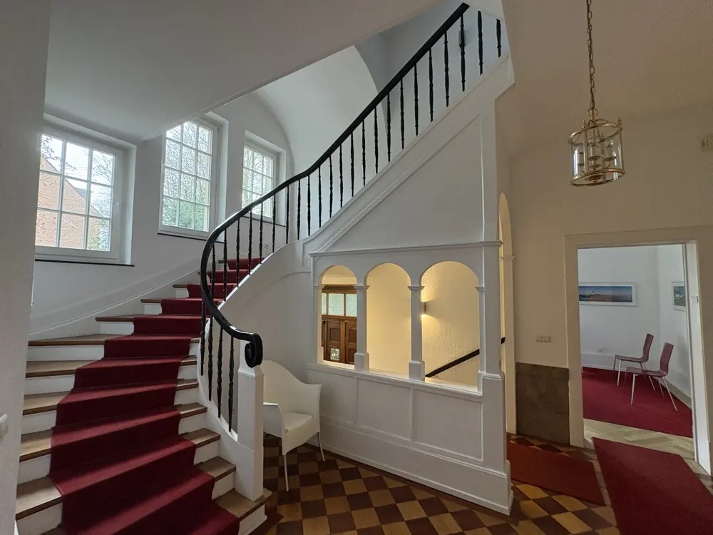 Interior view of a foyer with a winding staircase, red carpet, white walls, and checkered floor. A doorway leads to a room with red carpet and chairs.