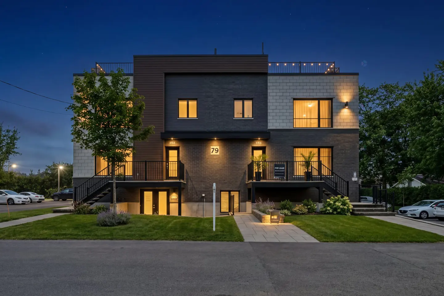 Modern townhouse at dusk with gray brick, brown siding, and black railings. Number 79 above the door. Rooftop lights visible.