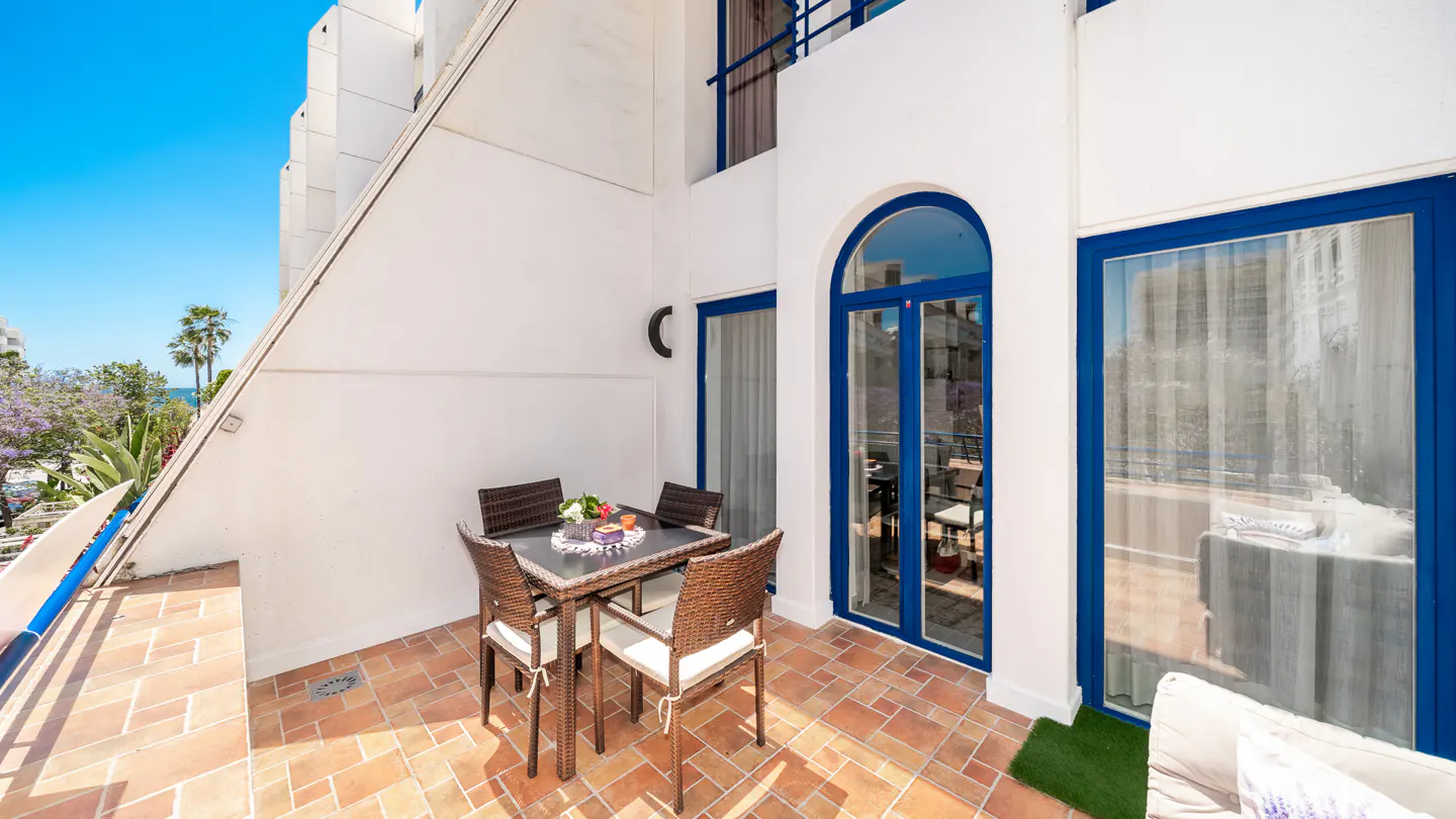 Outdoor patio with brown wicker table and chairs on a terracotta tiled floor. White walls with blue framed windows and doors. Ocean view in the background.