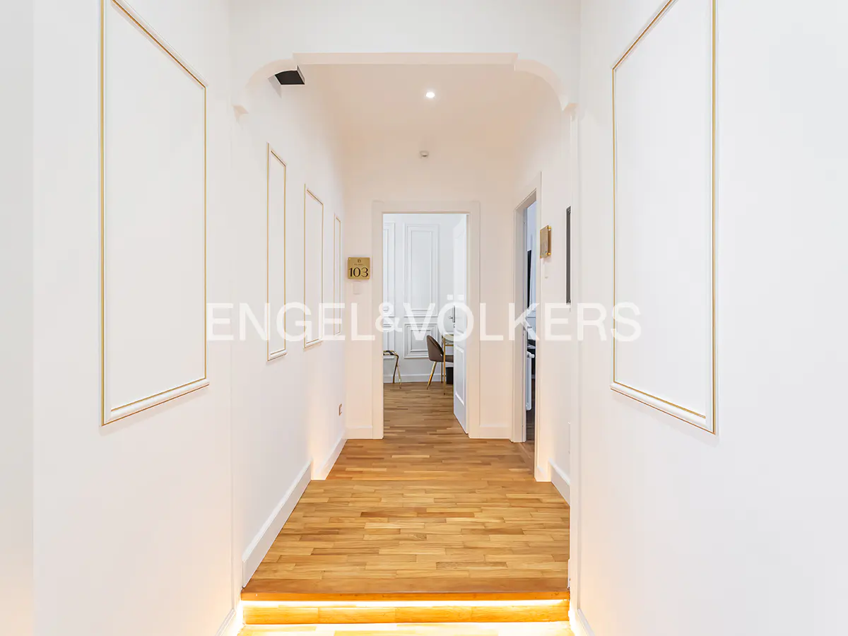Bright hallway with white walls, wood floors, and framed art. A doorway leads to a room with a desk and chair.
