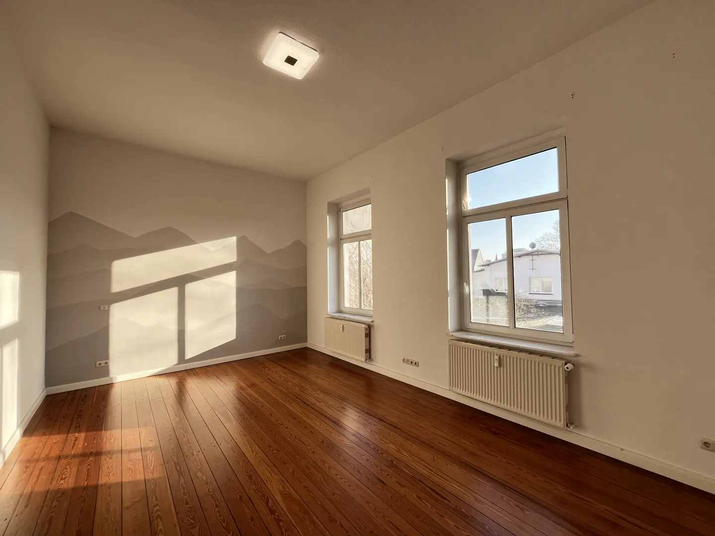 Bright, empty room with hardwood floors, white walls, and two windows. A gray mountain mural is on one wall.