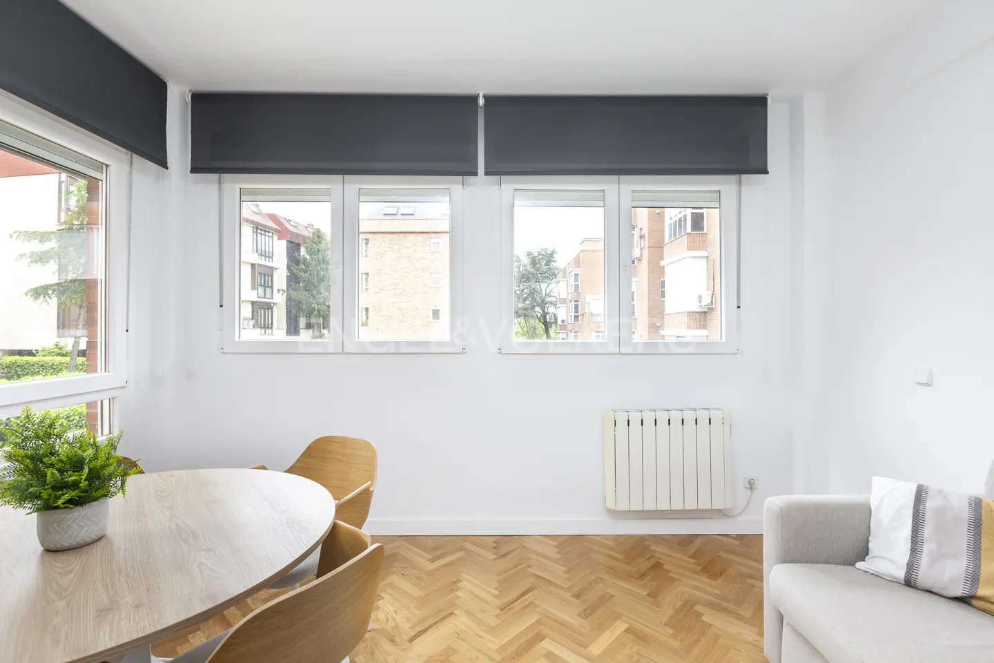 Bright living room with white walls, herringbone wood floor, round table, and gray sofa. Windows with black blinds overlook city buildings.