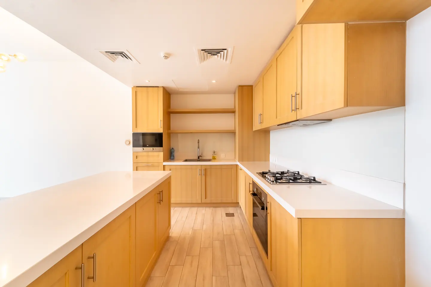 A bright kitchen with light wood cabinets, white countertops, and wood floors. A gas stovetop and oven are visible.