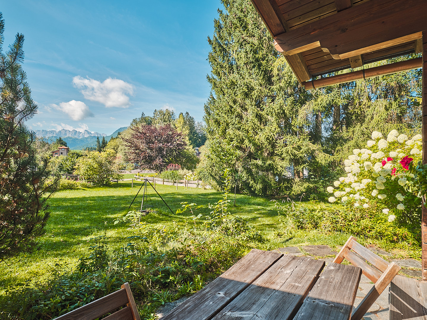 View from a wooden porch with table and chairs overlooking a green lawn, trees, and distant mountains under a blue sky. [AI-generated text]
