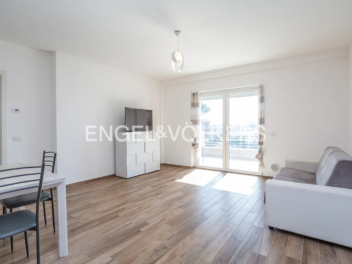 Bright living room with wood floors, white walls, and a sliding glass door to a balcony. A gray sofa and white table with chairs are visible.