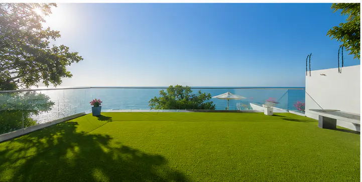Balcony with green turf, glass railing, and ocean view. A white umbrella and potted flowers add to the serene scene.