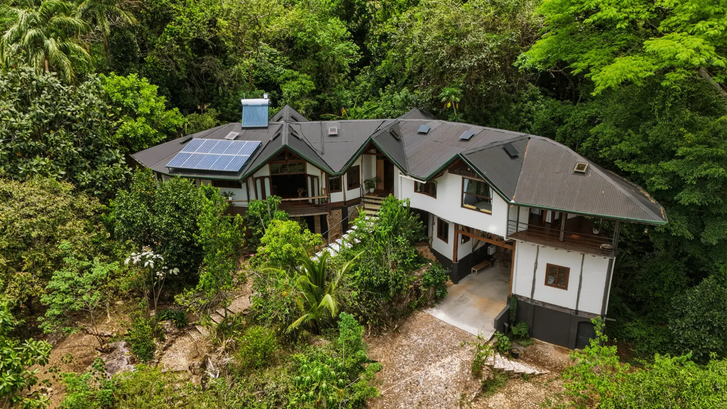 Aerial view of a modern white house with a black roof, solar panels, and lush green trees surrounding it.