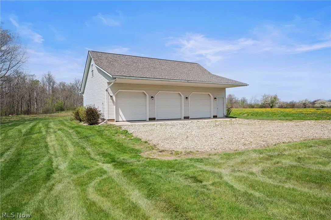 A three-car garage with white doors and beige siding sits on a gravel driveway surrounded by green grass and a field of yellow flowers.