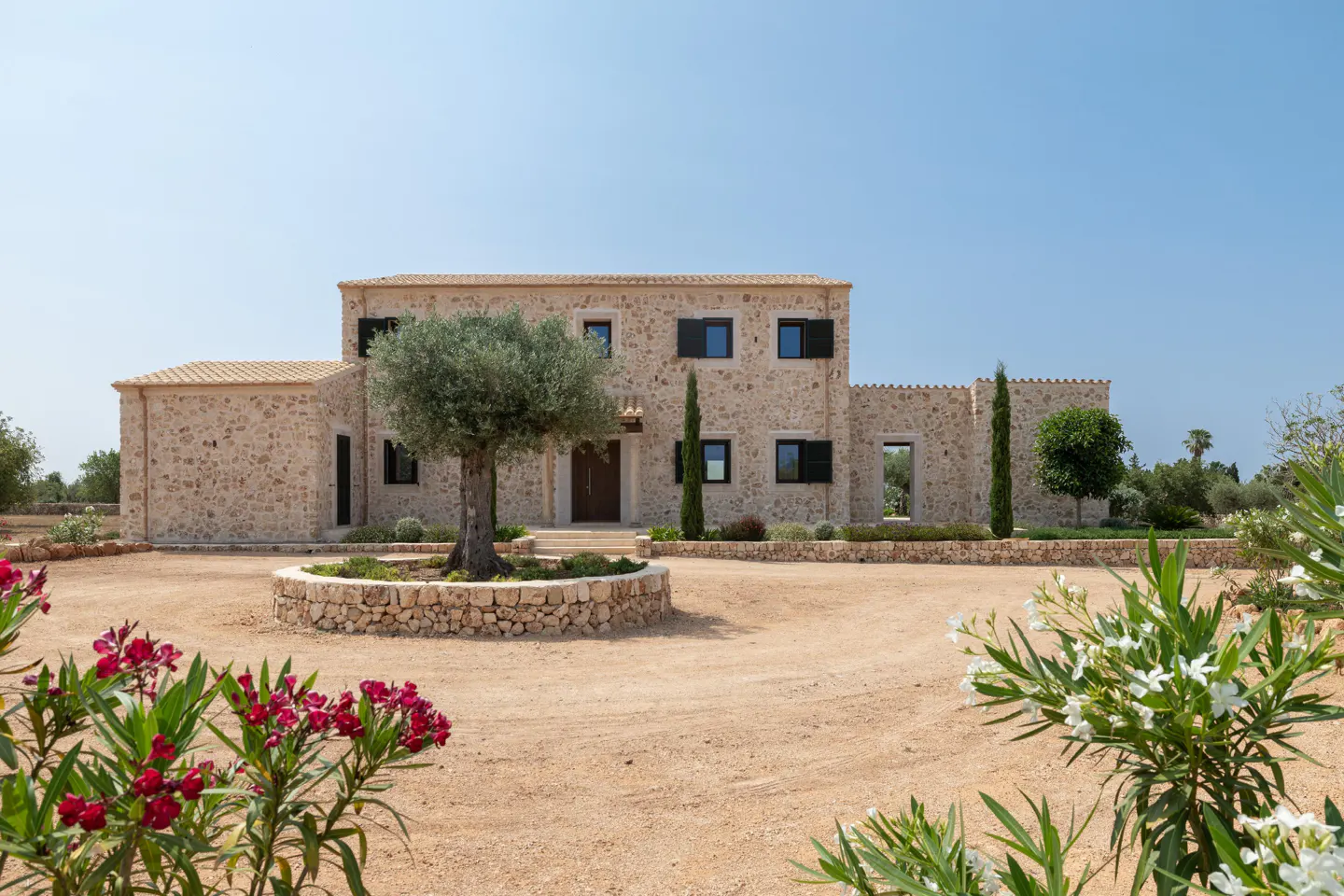 Two-story stone house with black shutters, a brown door, and a tree in the front yard. Red and white flowers are in the foreground.