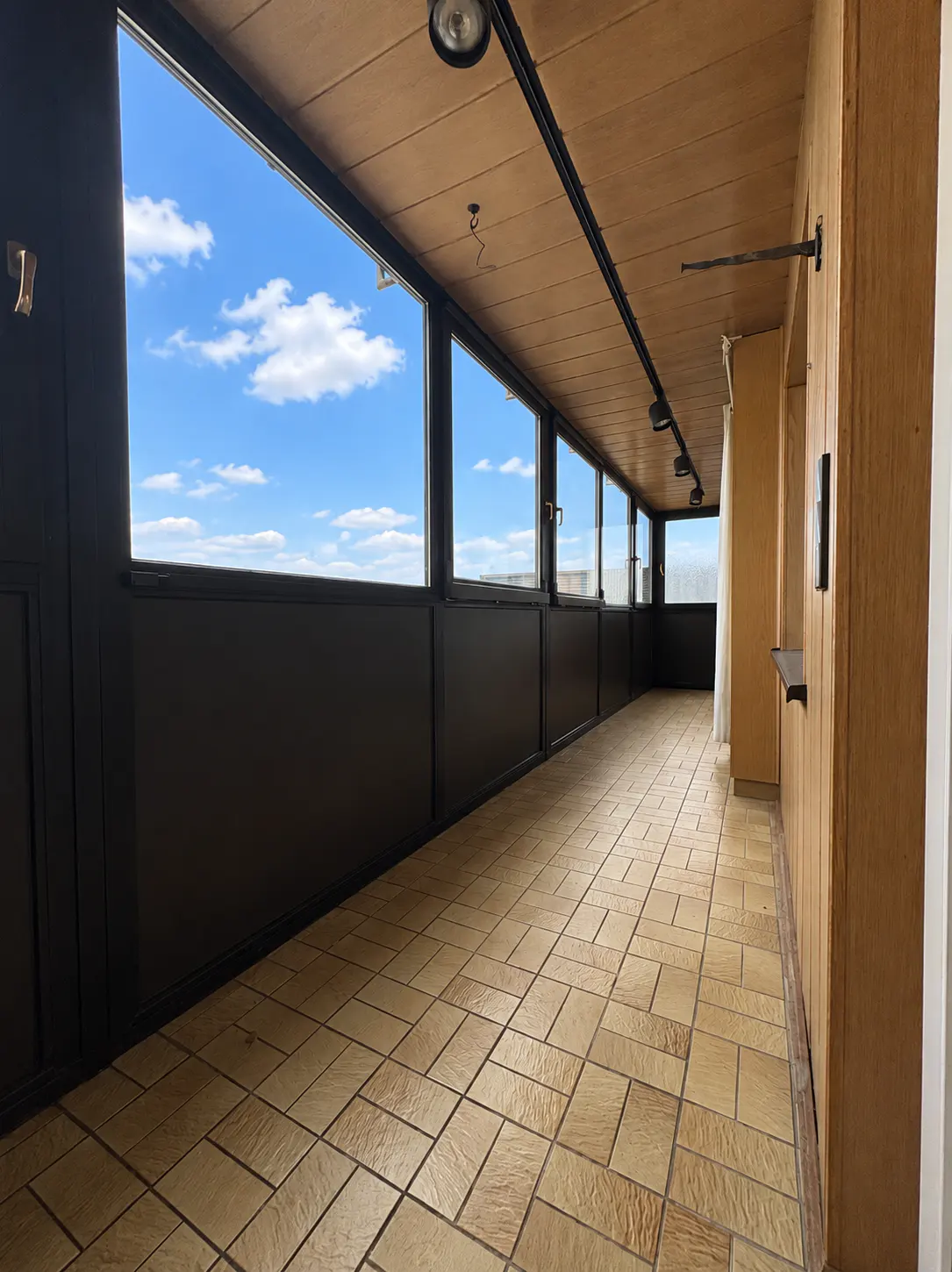 A long, narrow room with tan tile floors, dark walls with windows, and a wood paneled wall.