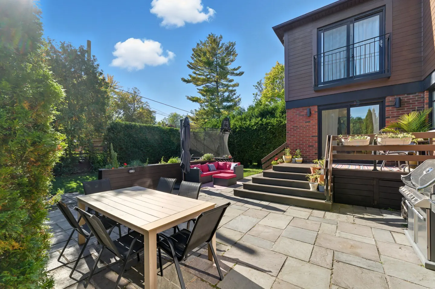 Backyard patio with stone pavers, dining table, red outdoor sofa, wooden deck with stairs, and a grill under a blue sky.