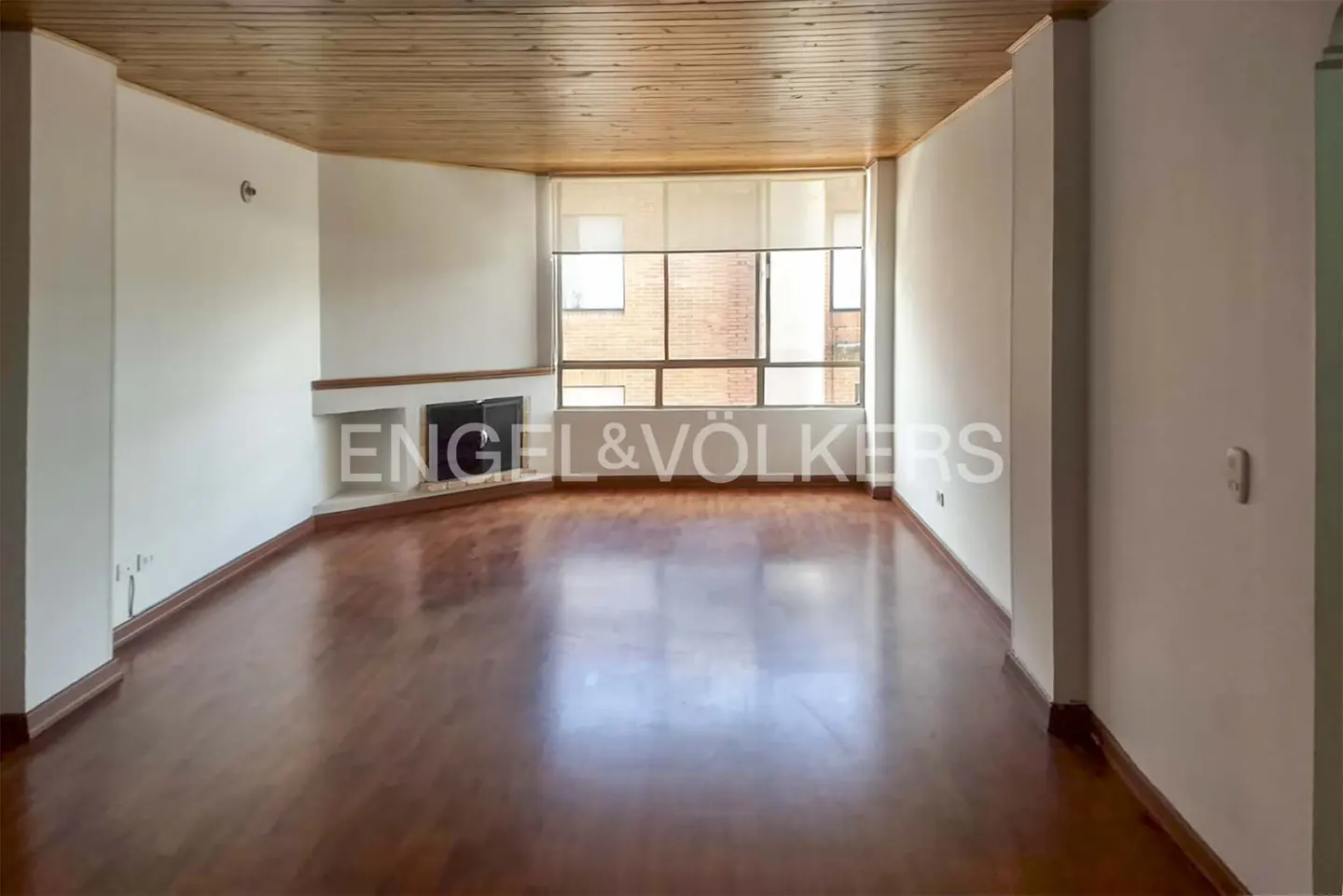Empty living room with hardwood floors, a fireplace, and a large window with a view of a brick building. The ceiling is wood.