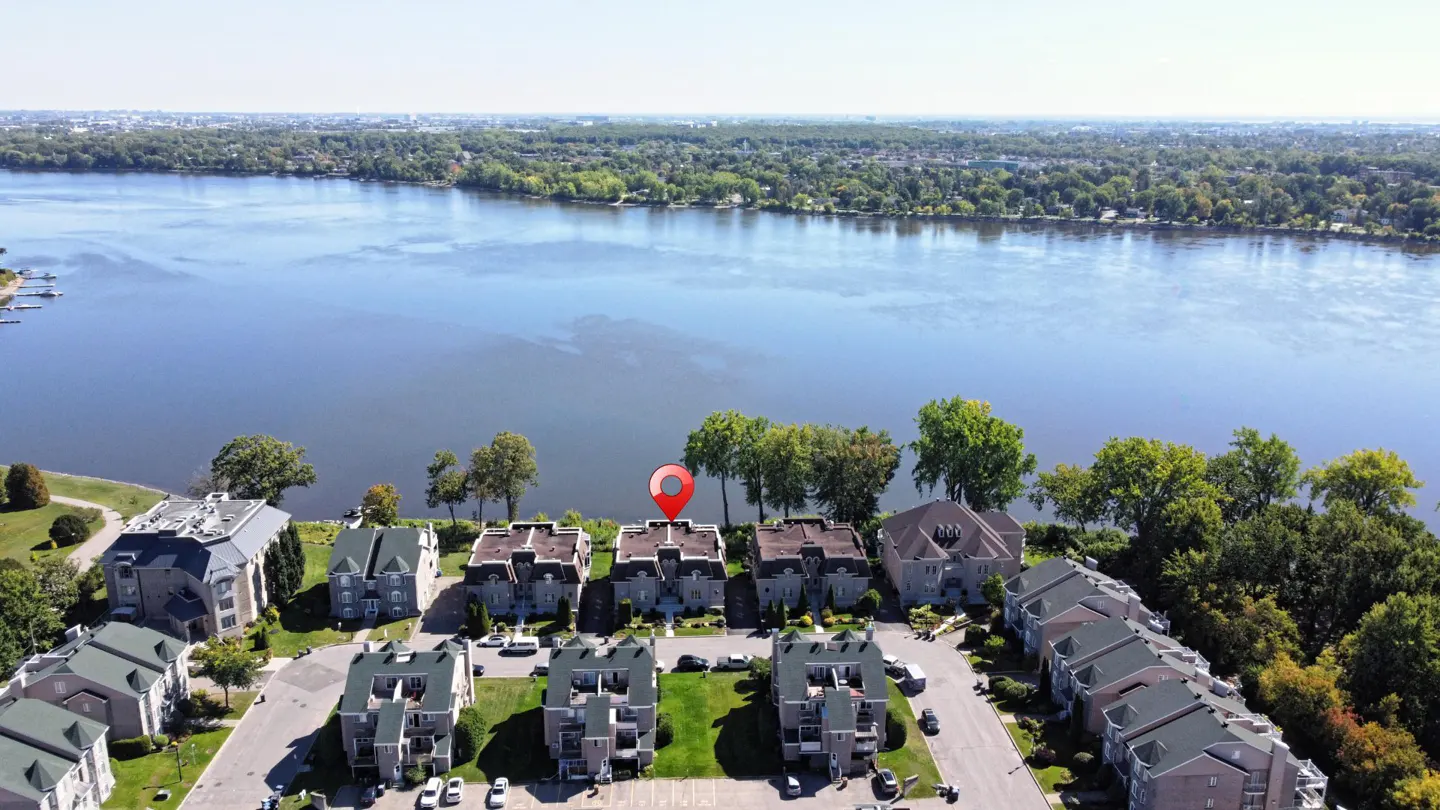 Aerial view of gray townhouses with a red location pin on the roof, near a blue river and green trees.