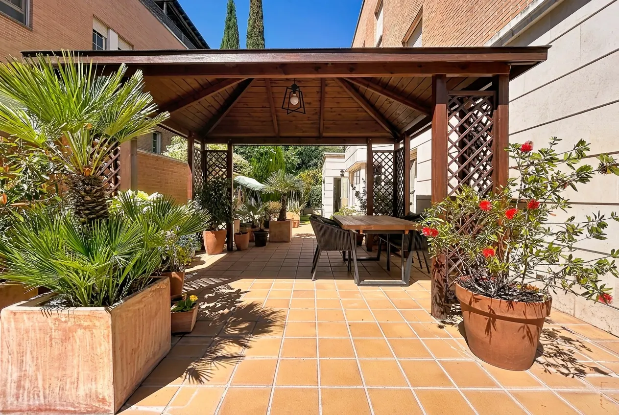 Outdoor patio with a wooden gazebo, table, chairs, and potted plants on a tiled floor.