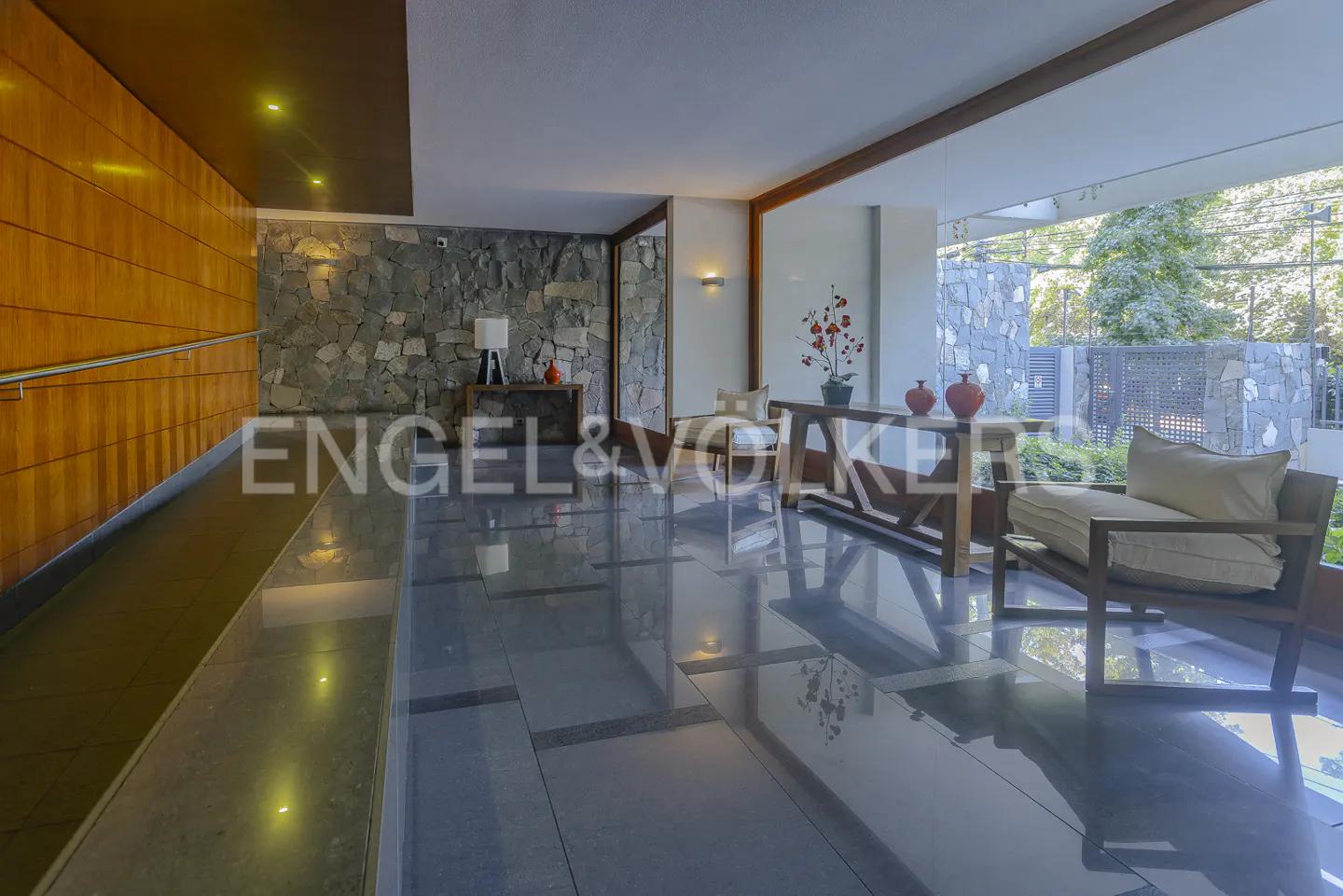 Modern lobby with stone walls, wood accents, and polished floors. A chair and table sit near a window overlooking greenery.