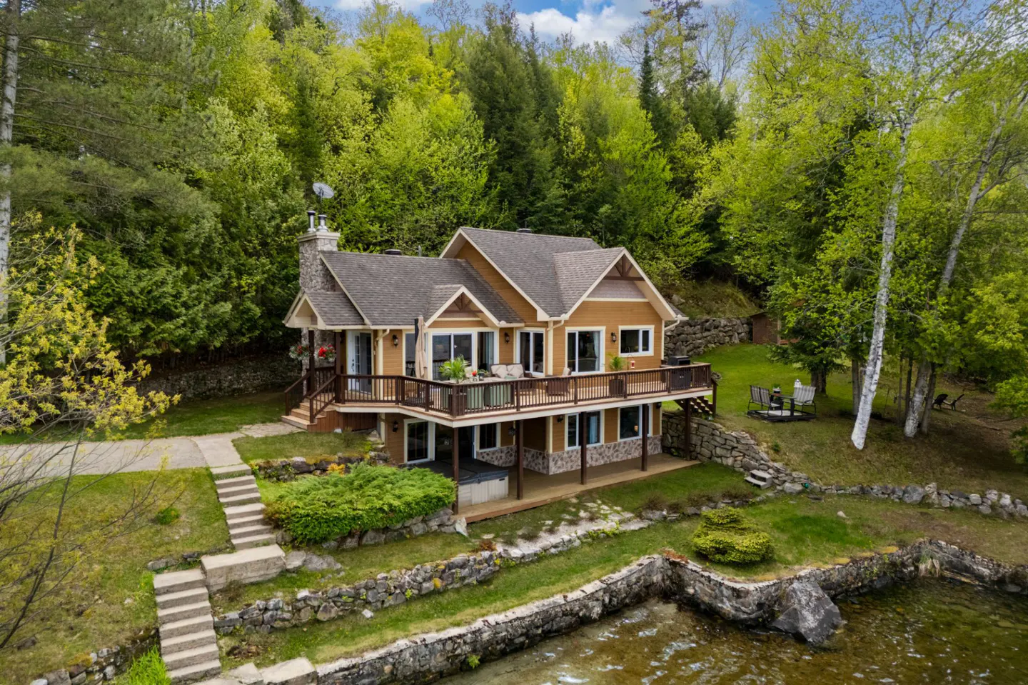 Two-story tan house with a large deck, stone chimney, and stone retaining wall, nestled in a lush green forest by a lake.