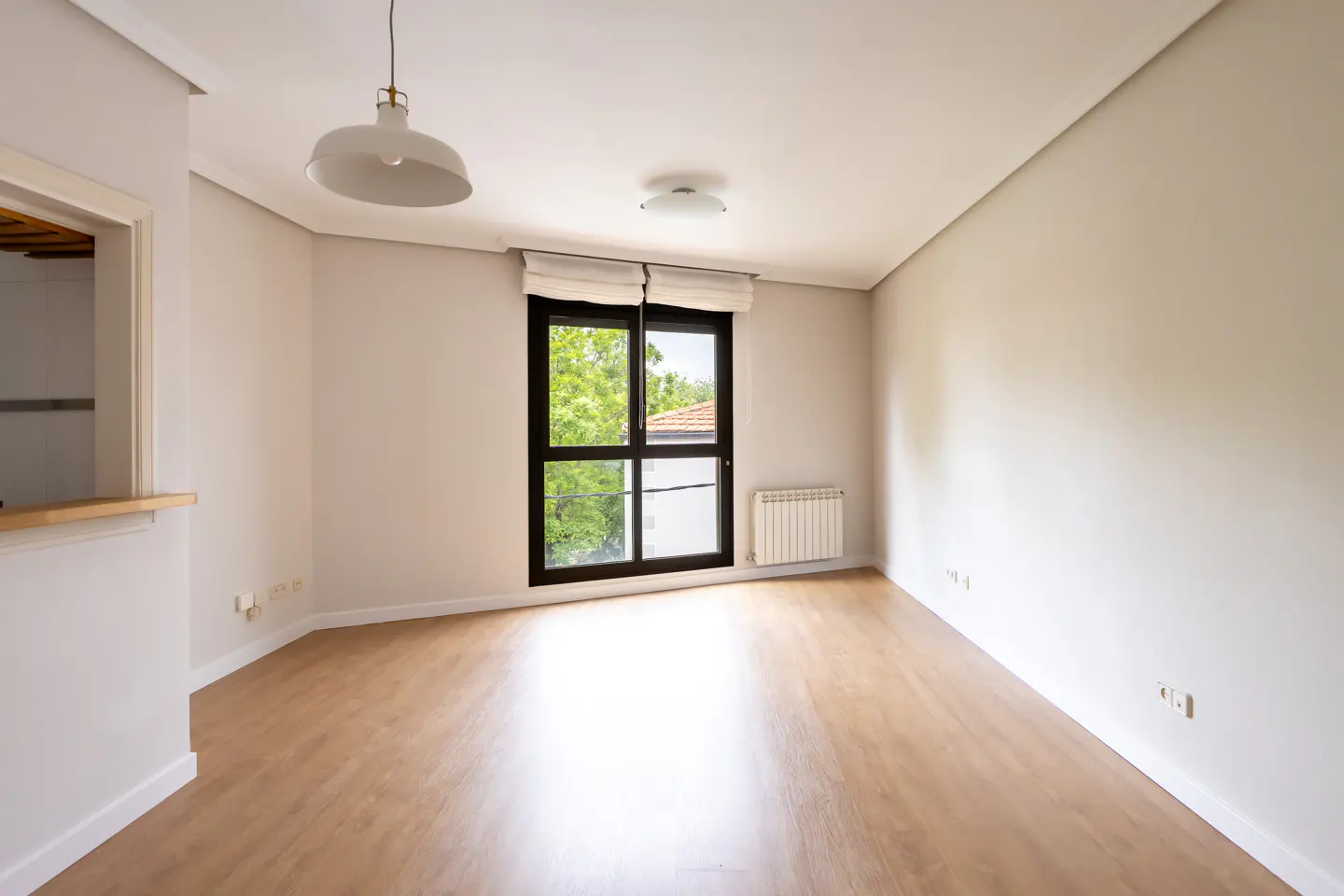 Bright, empty room with light wood floors, white walls, and a black-framed window with green trees visible outside. A white pendant light hangs from the ceiling.