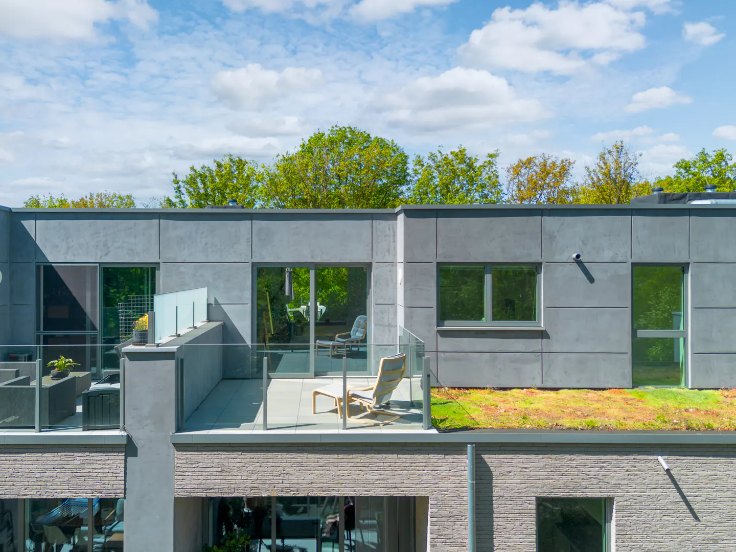 Modern gray building with balconies and a green roof under a blue sky with white clouds. Balconies have glass railings and outdoor furniture.