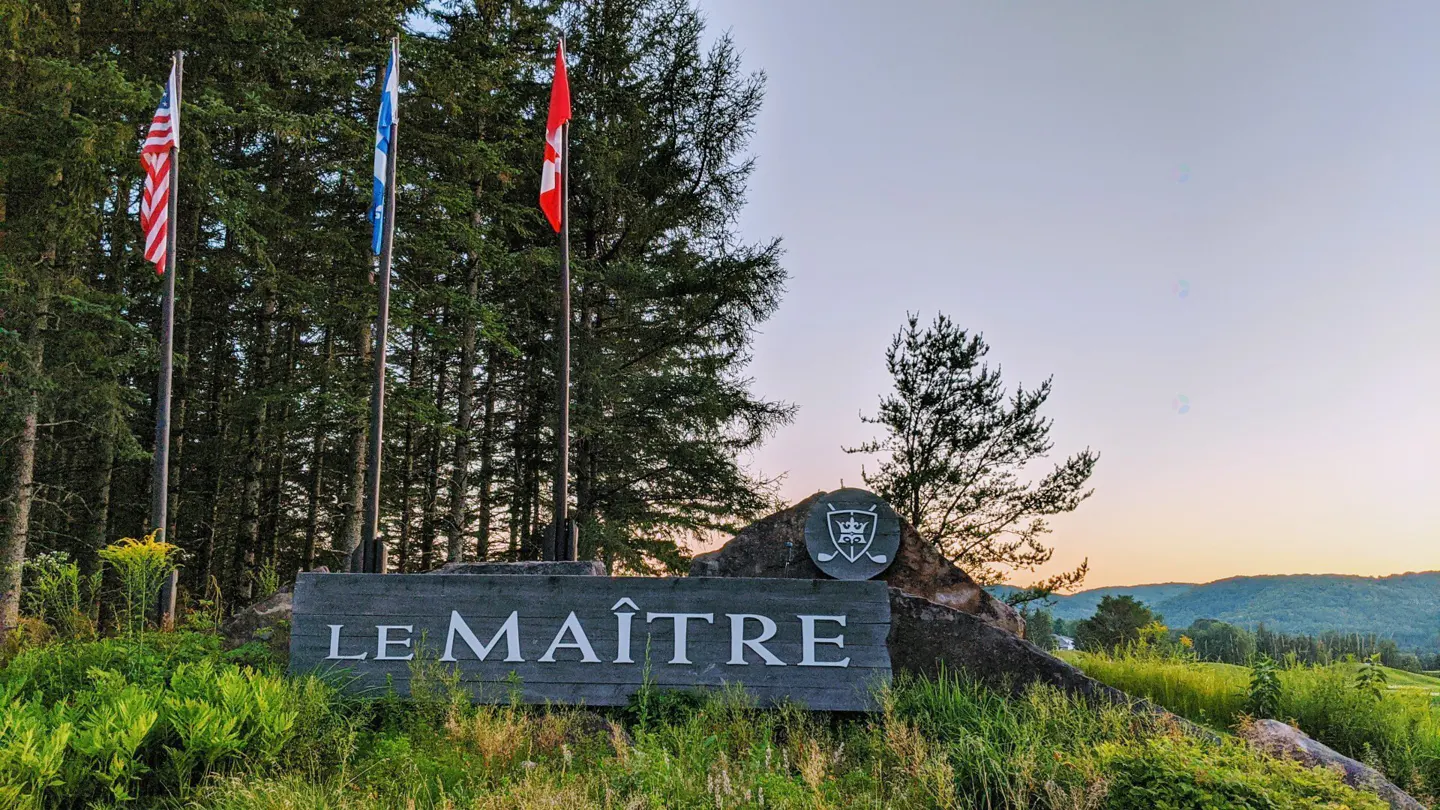 Entrance to Le Maître golf course with US, Quebec, and Canadian flags. Sign is gray stone with white letters. Trees and hills in background.