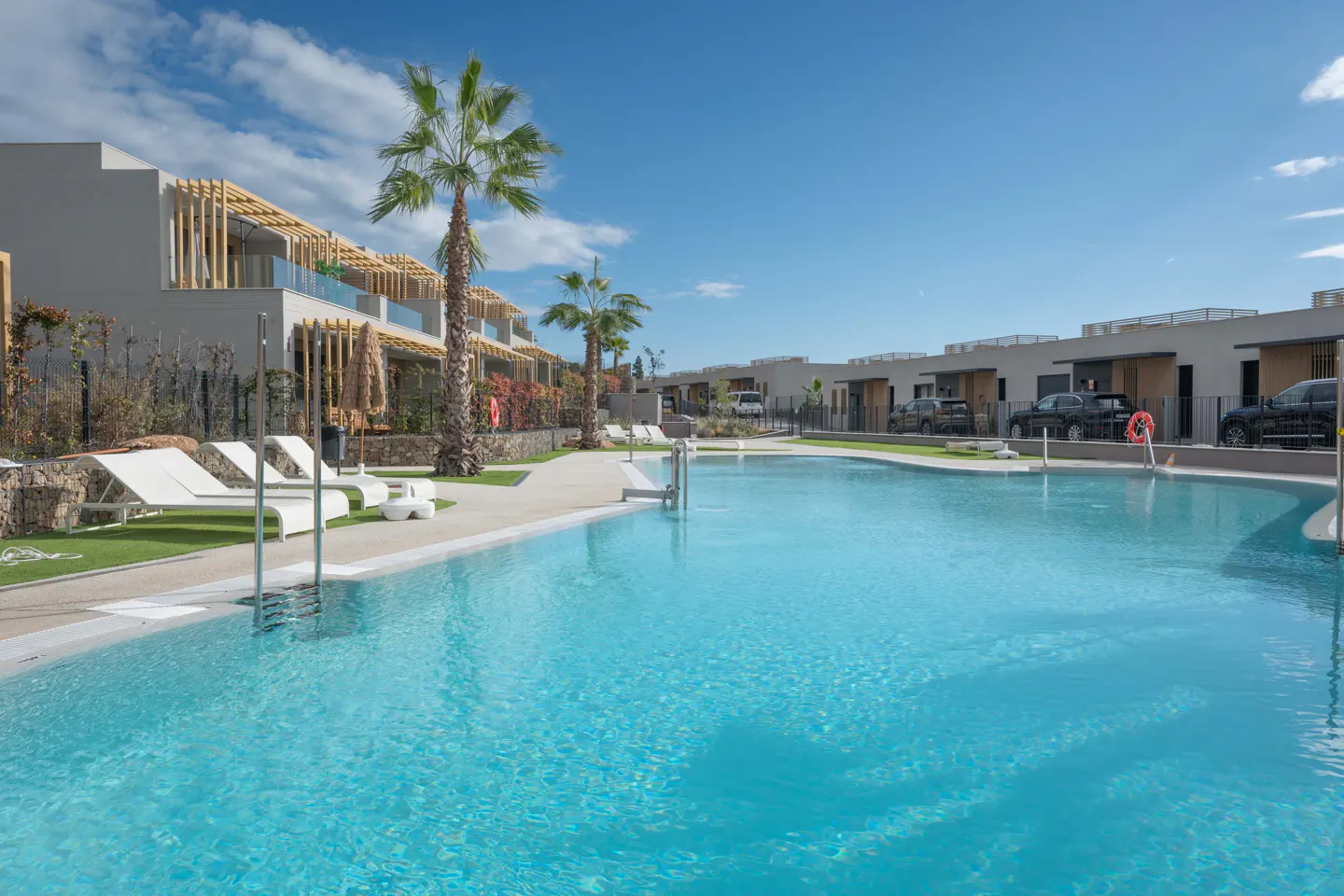 A wide shot of a turquoise pool surrounded by modern apartments and palm trees under a blue sky.
